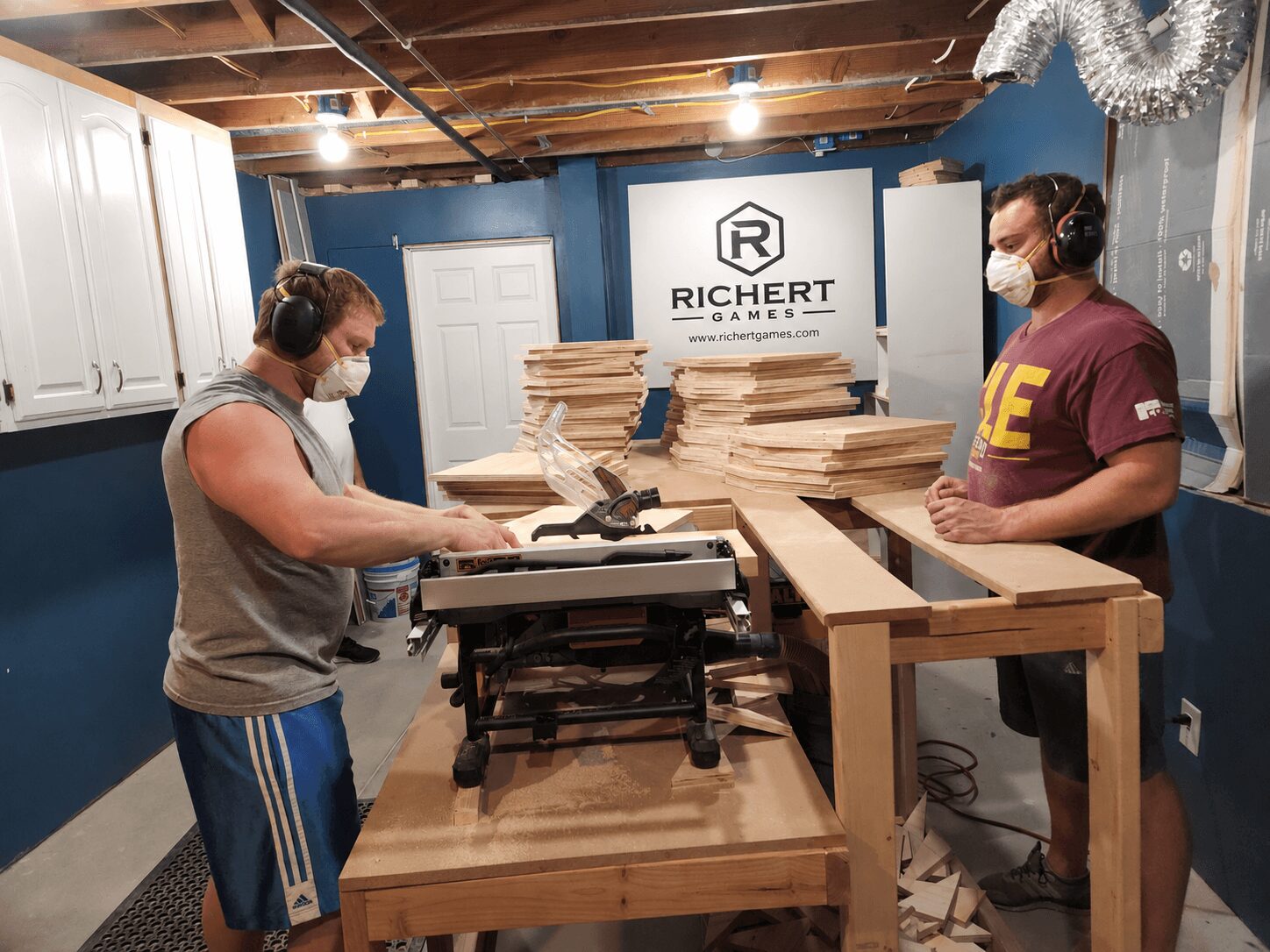 Two people wearing masks work with paper stacks and a machine in a room with blue walls and wooden ceiling beams.