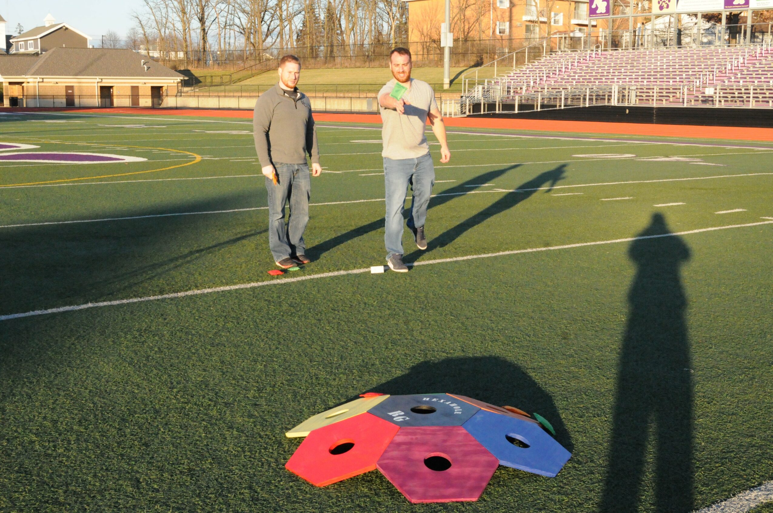 Two people walking on a sports field with a colorful game piece on the ground, shadows cast by the sun.