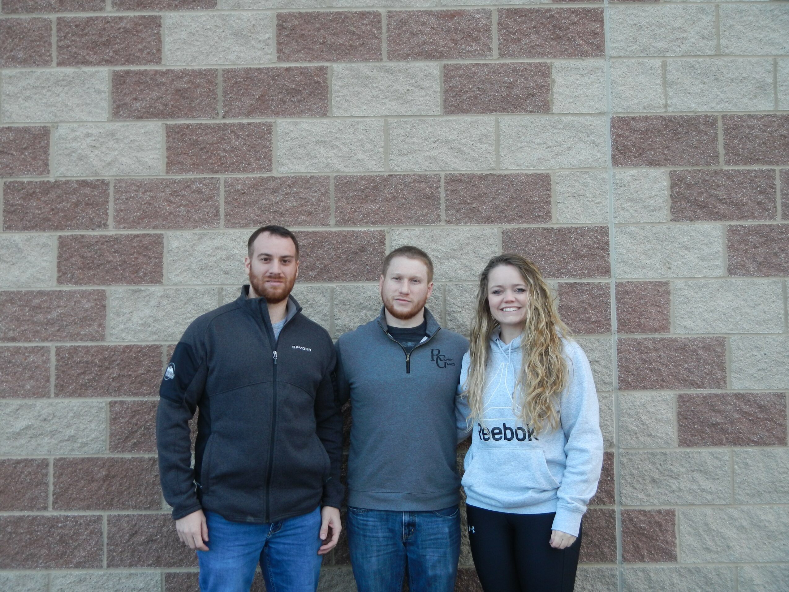 Three people standing in front of a brick wall, two men and one woman, smiling.