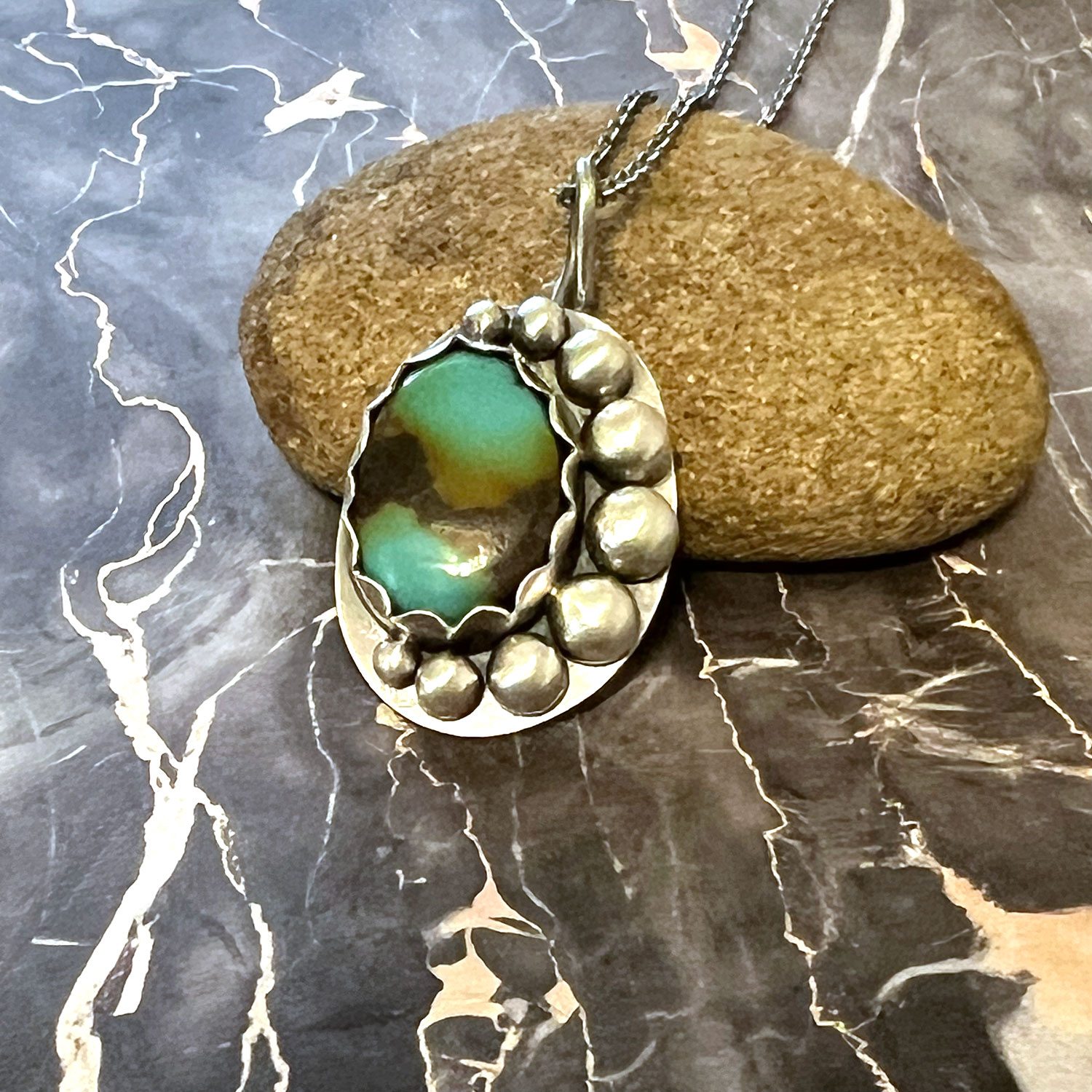 Pendant with a green and brown stone surrounded by silver beads, resting on a brown stone on a marble surface.