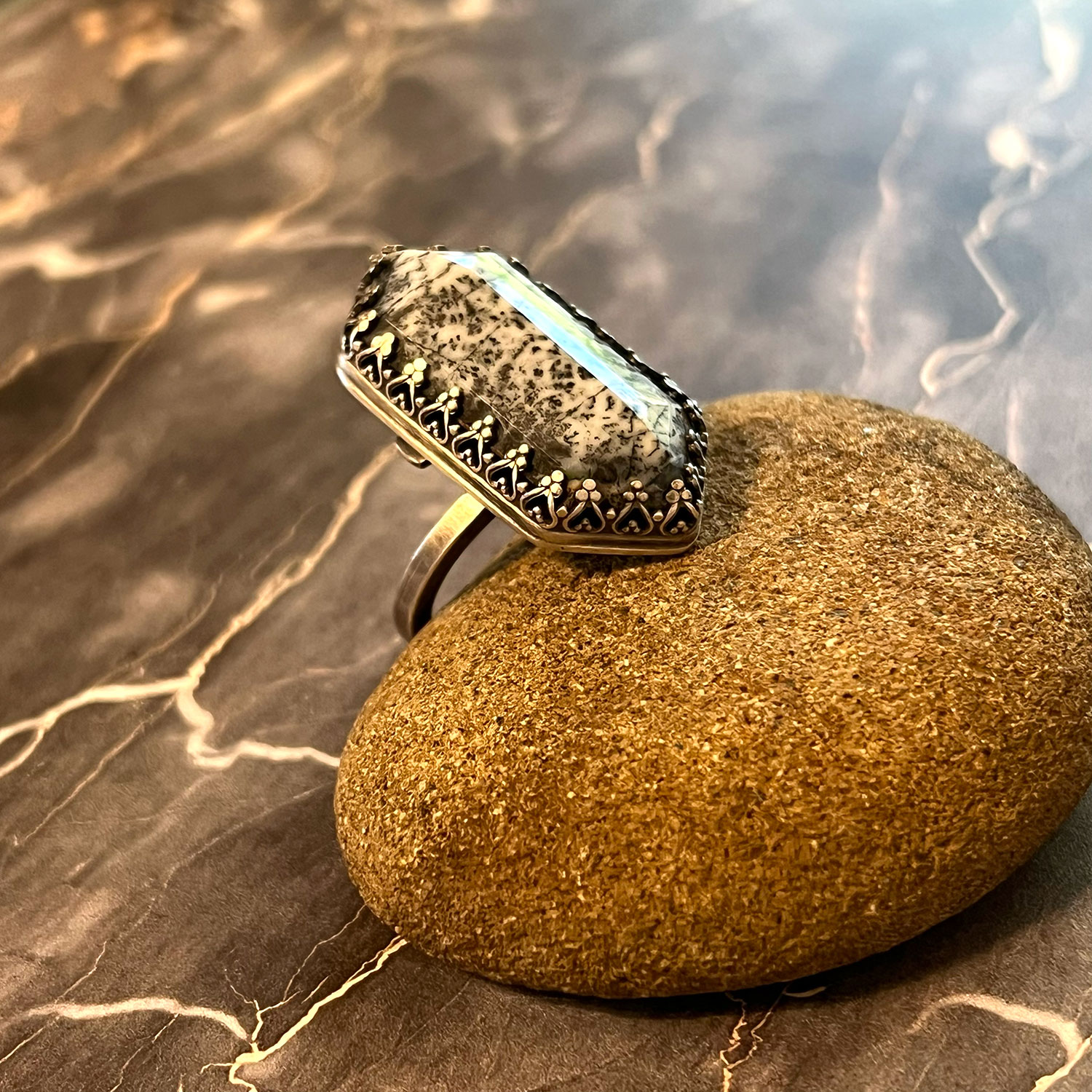 A ring with a large, oval-shaped stone rests on a smooth, rounded rock, with a textured surface and a reflective background.