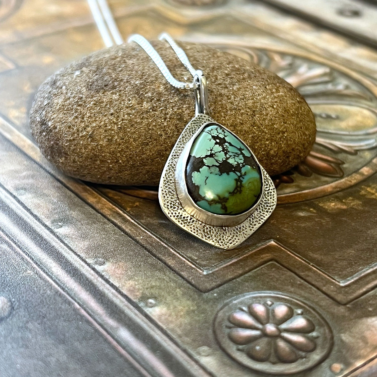 Silver pendant with turquoise stone resting on a smooth stone, on a metallic surface with floral engravings.