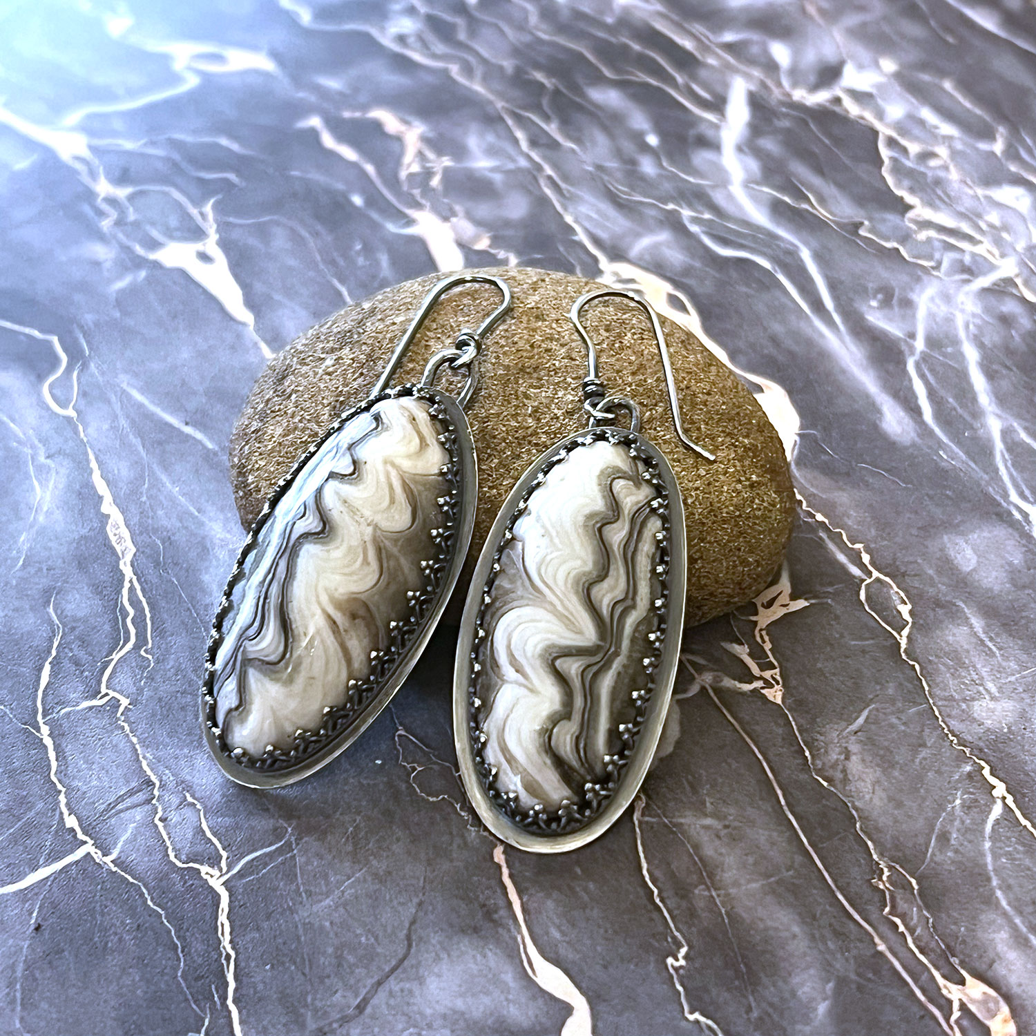 Oval-shaped earrings with black and white marbled pattern, hanging on a textured stone, on a marble surface background.