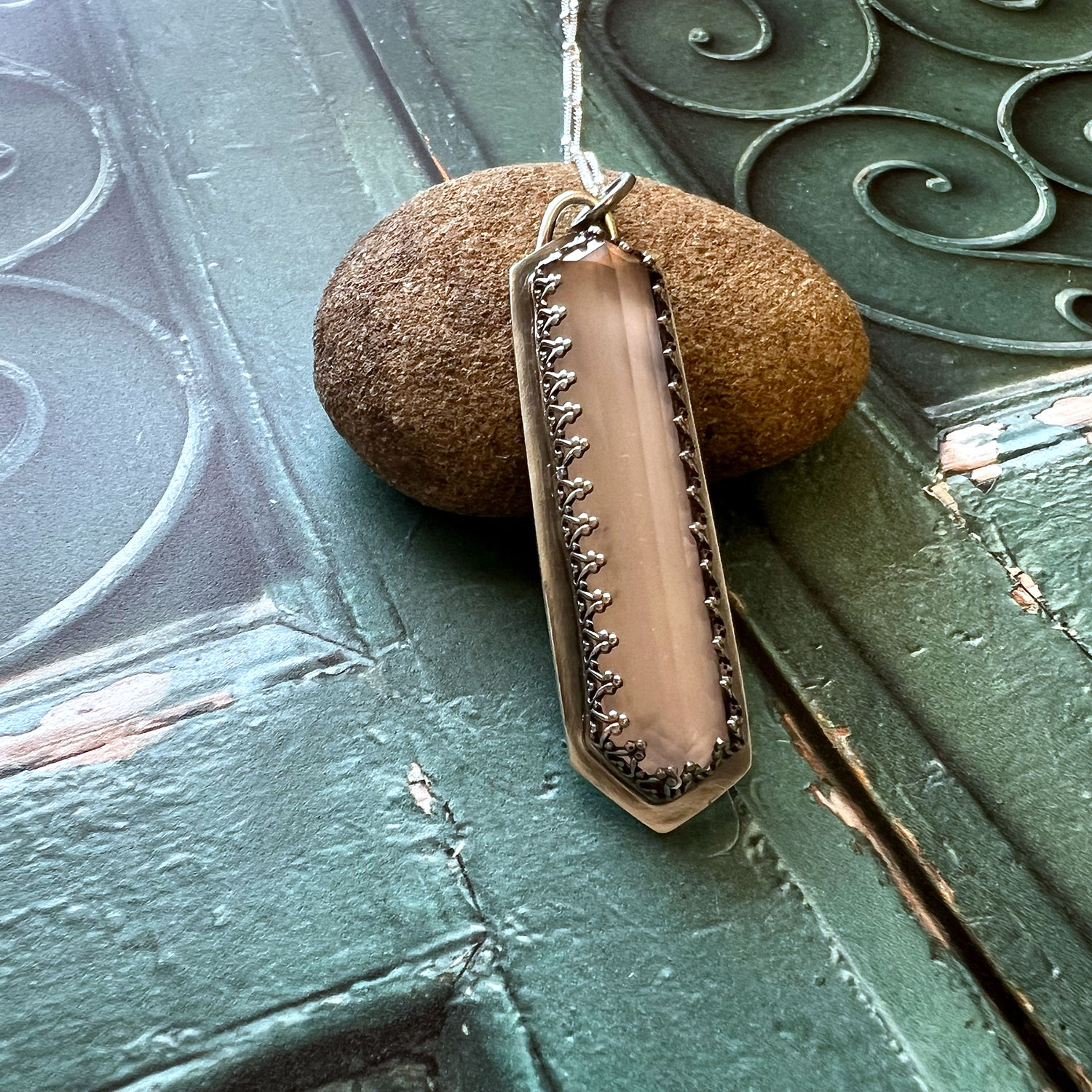 Pendant with a rectangular, beige stone surrounded by a metal frame, hanging on a chain, resting against a rock and green surface.