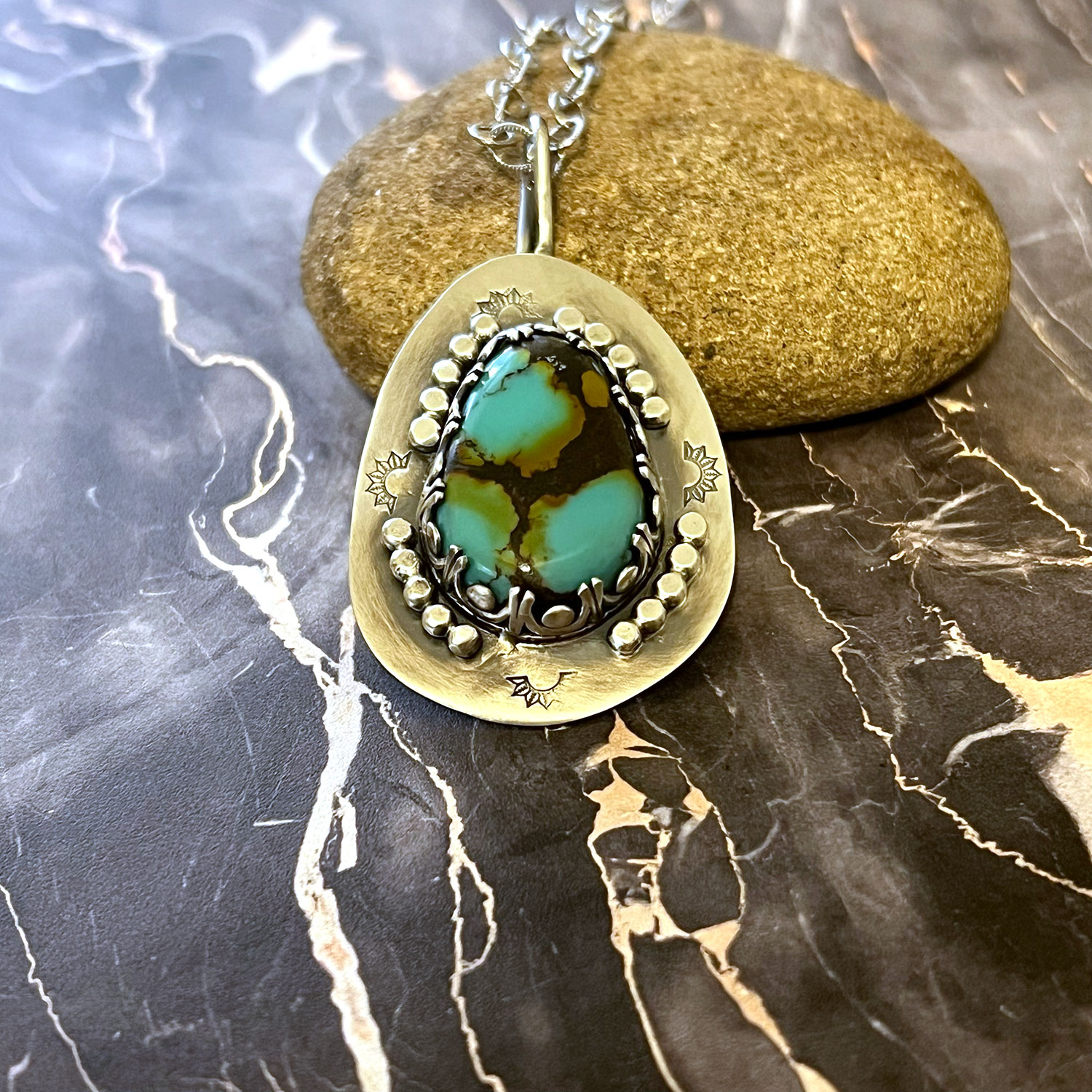 Pendant with turquoise and black stone, silver accents, hanging from a chain, placed on a rock and marble surface.
