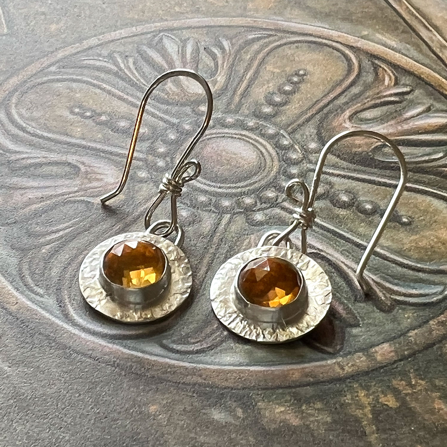 Pair of earrings with round amber-colored stones and textured silver discs, on a decorative surface.