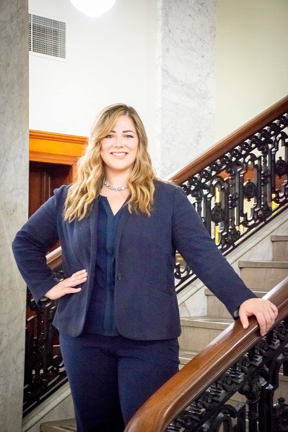 Woman with blonde hair smiling, standing on a staircase with a wooden handrail, wearing a dark blazer and pants.