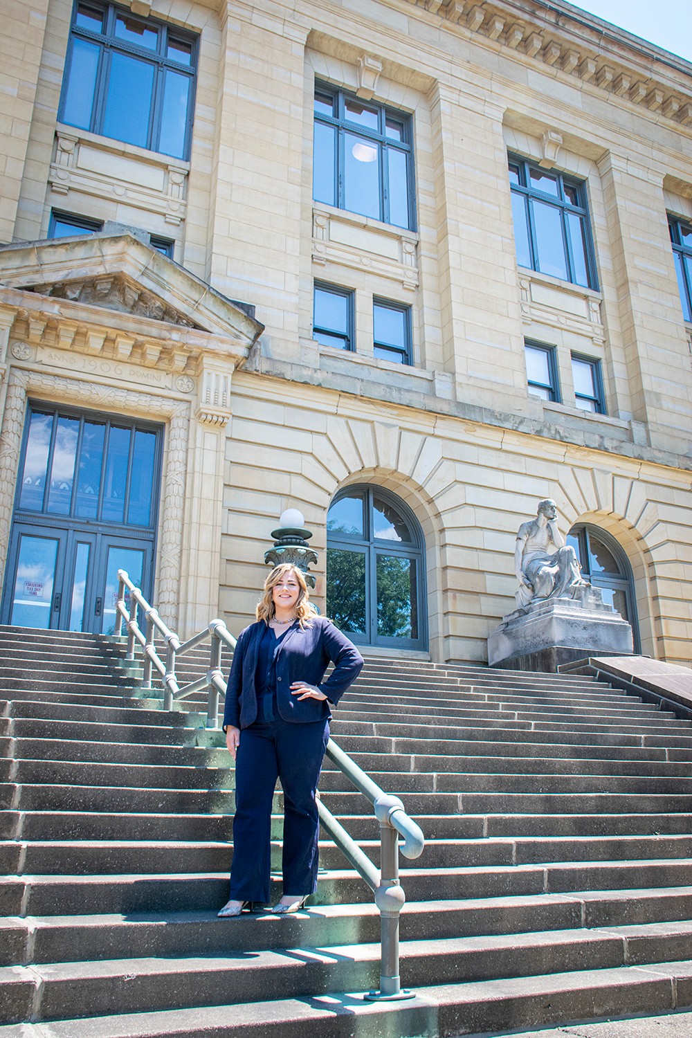 Woman in navy outfit standing on stairs outside a historic building with large windows and statues, smiling.