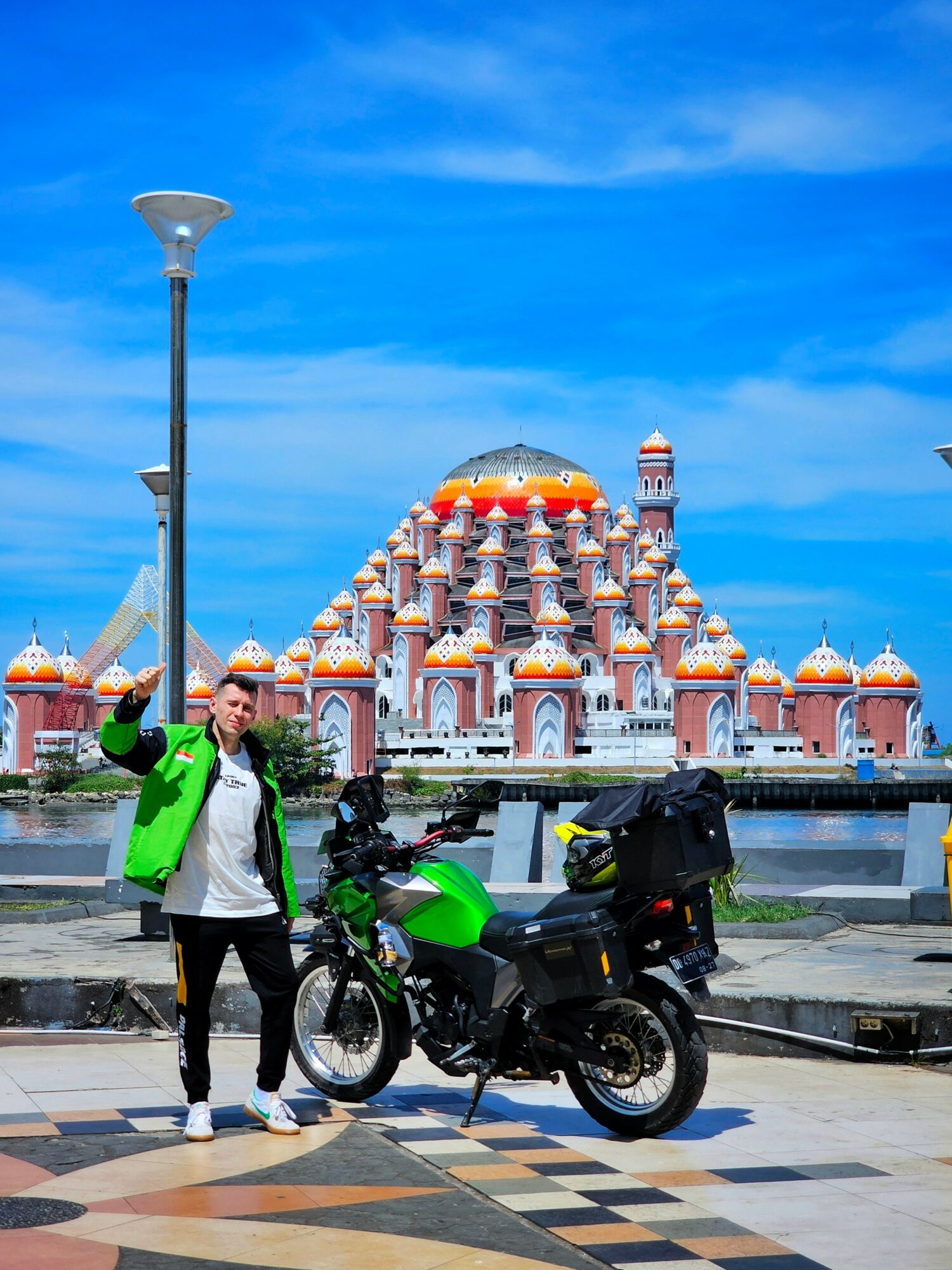 Person in green jacket standing next to a green motorcycle with a large building in the background under a blue sky.