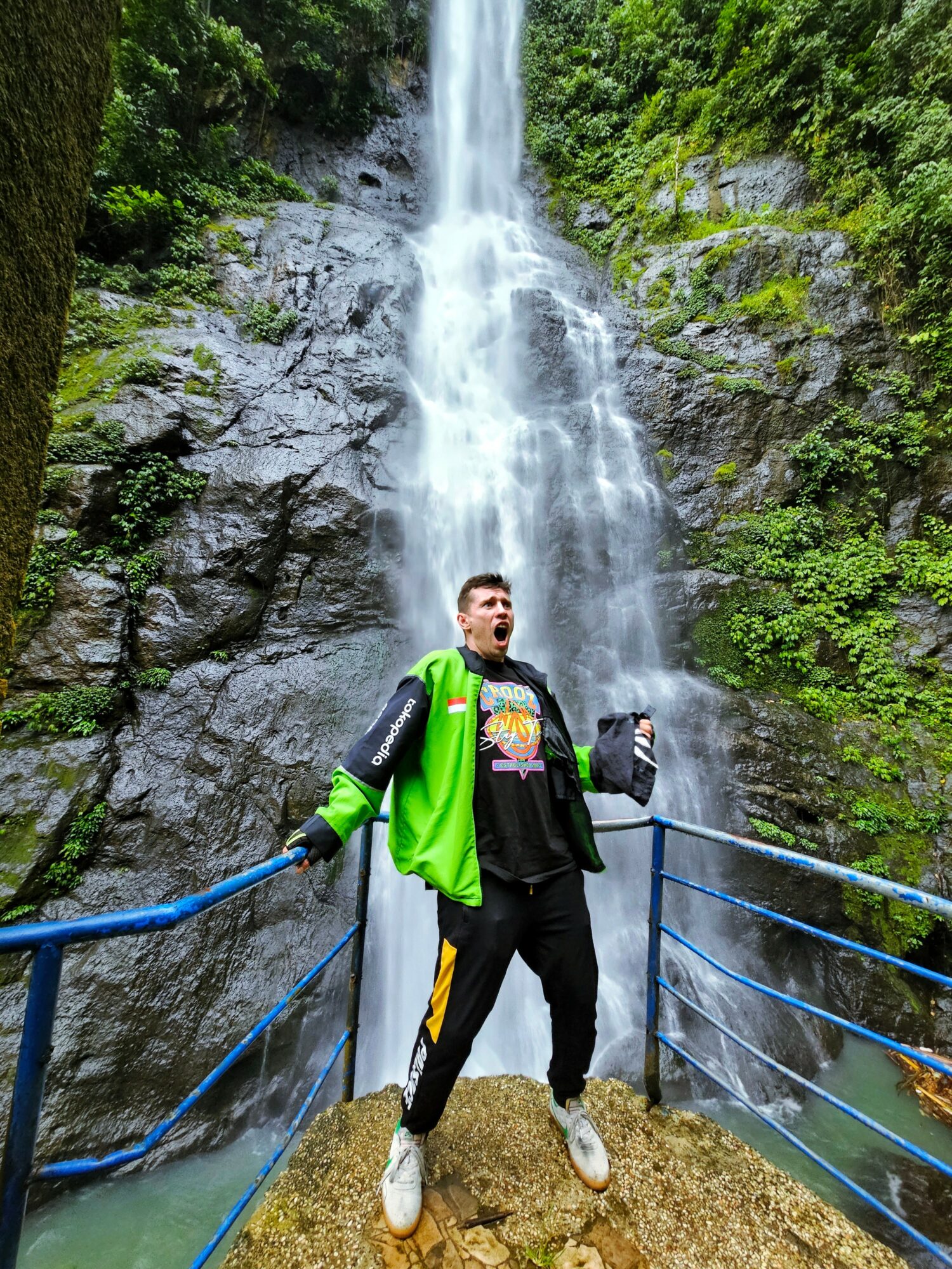 Person standing on a bridge in front of a tall waterfall surrounded by green foliage, holding a drink, wearing a green jacket and black pants.