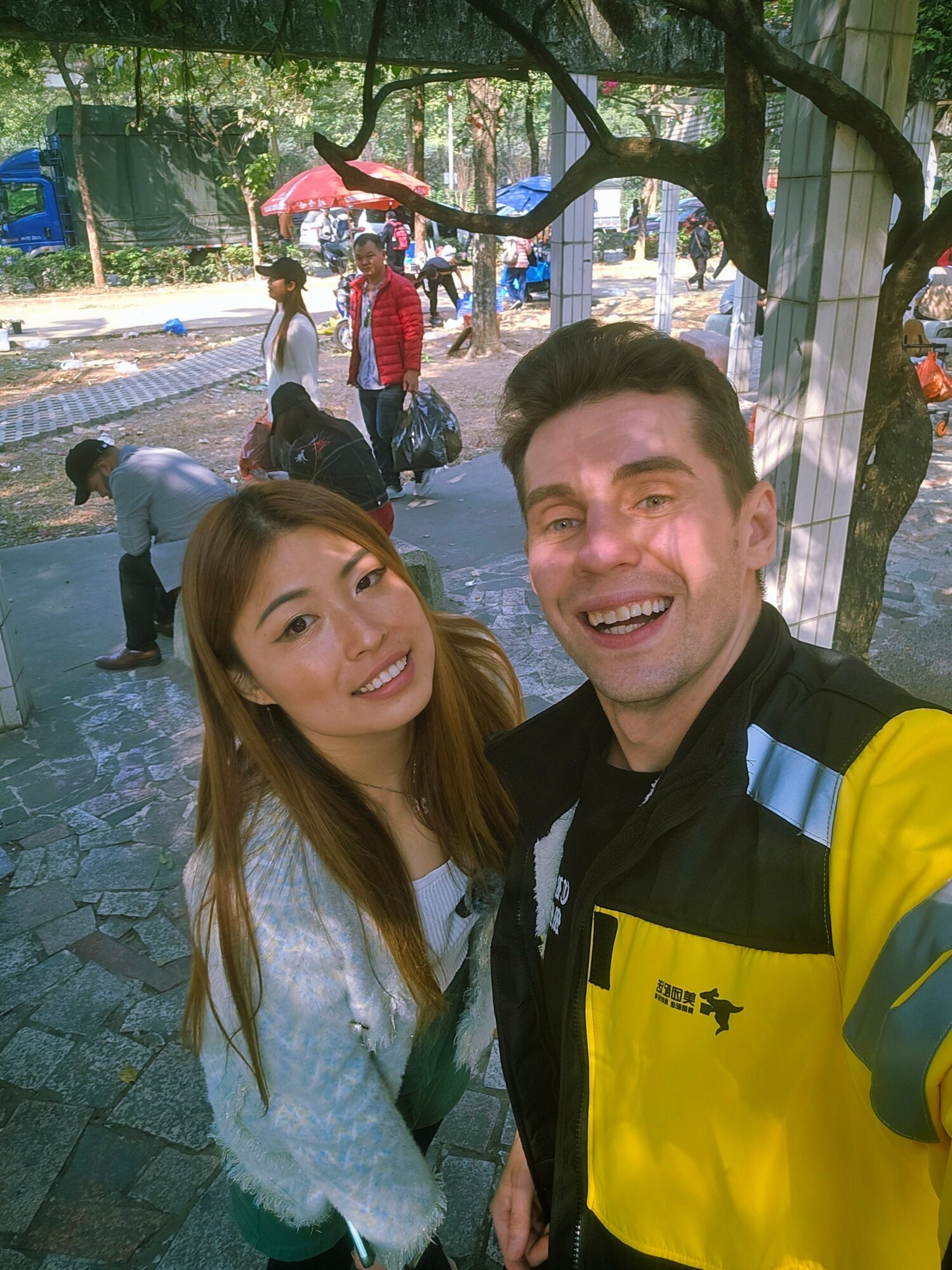 Two people smiling outdoors under a tree, with a park and other people in the background.