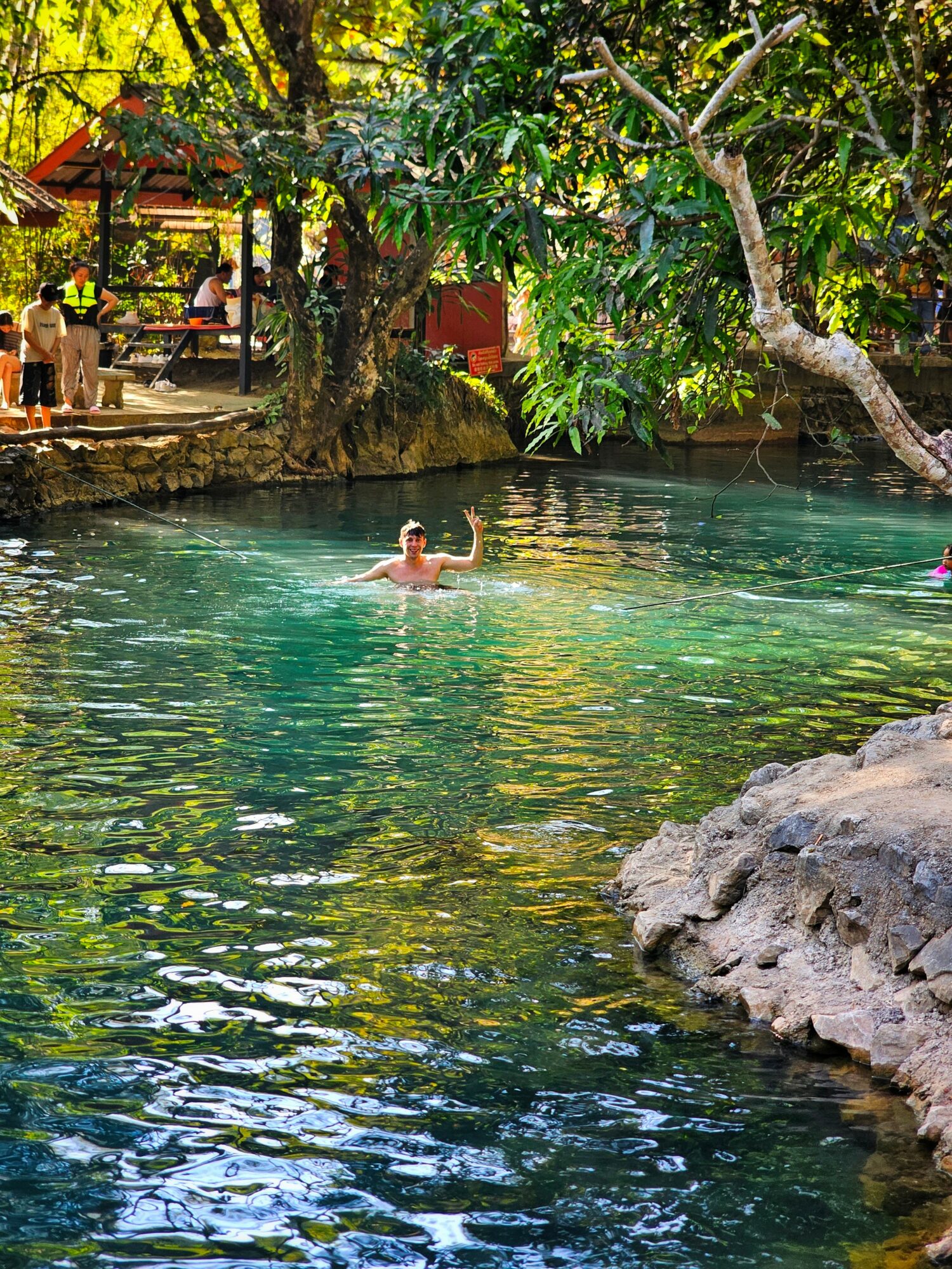 Person swimming in a clear, greenish-blue natural pool surrounded by trees and rocks, with a pavilion and people in the background.
