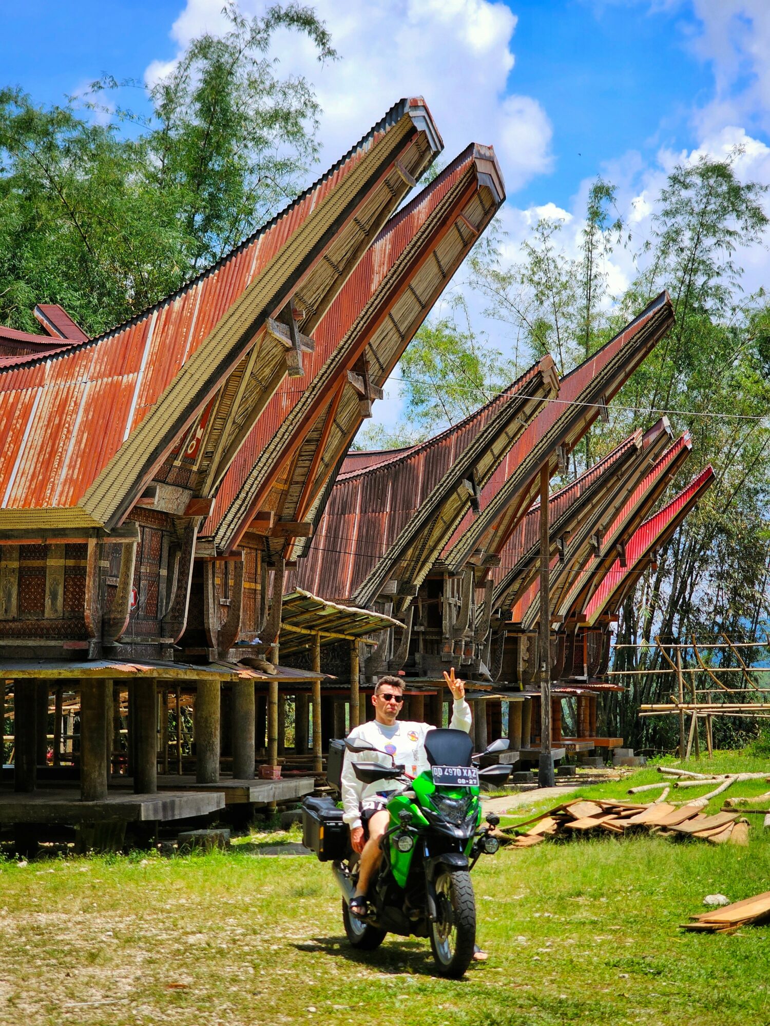 Person sitting on a green motorcycle in front of traditional wooden houses with curved roofs, trees, and a blue sky.