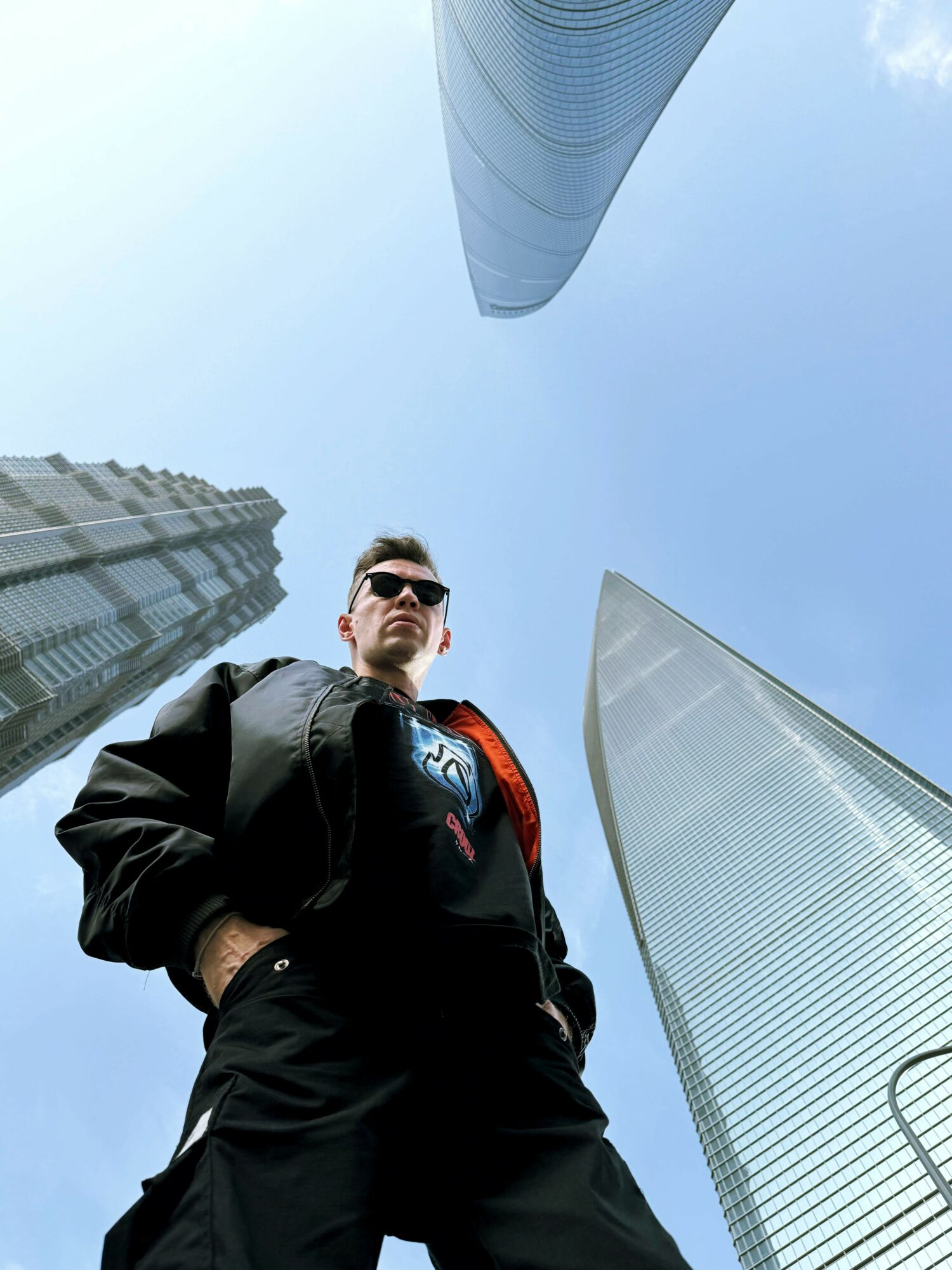 Low-angle view of a man in sunglasses standing among tall skyscrapers against a clear blue sky.