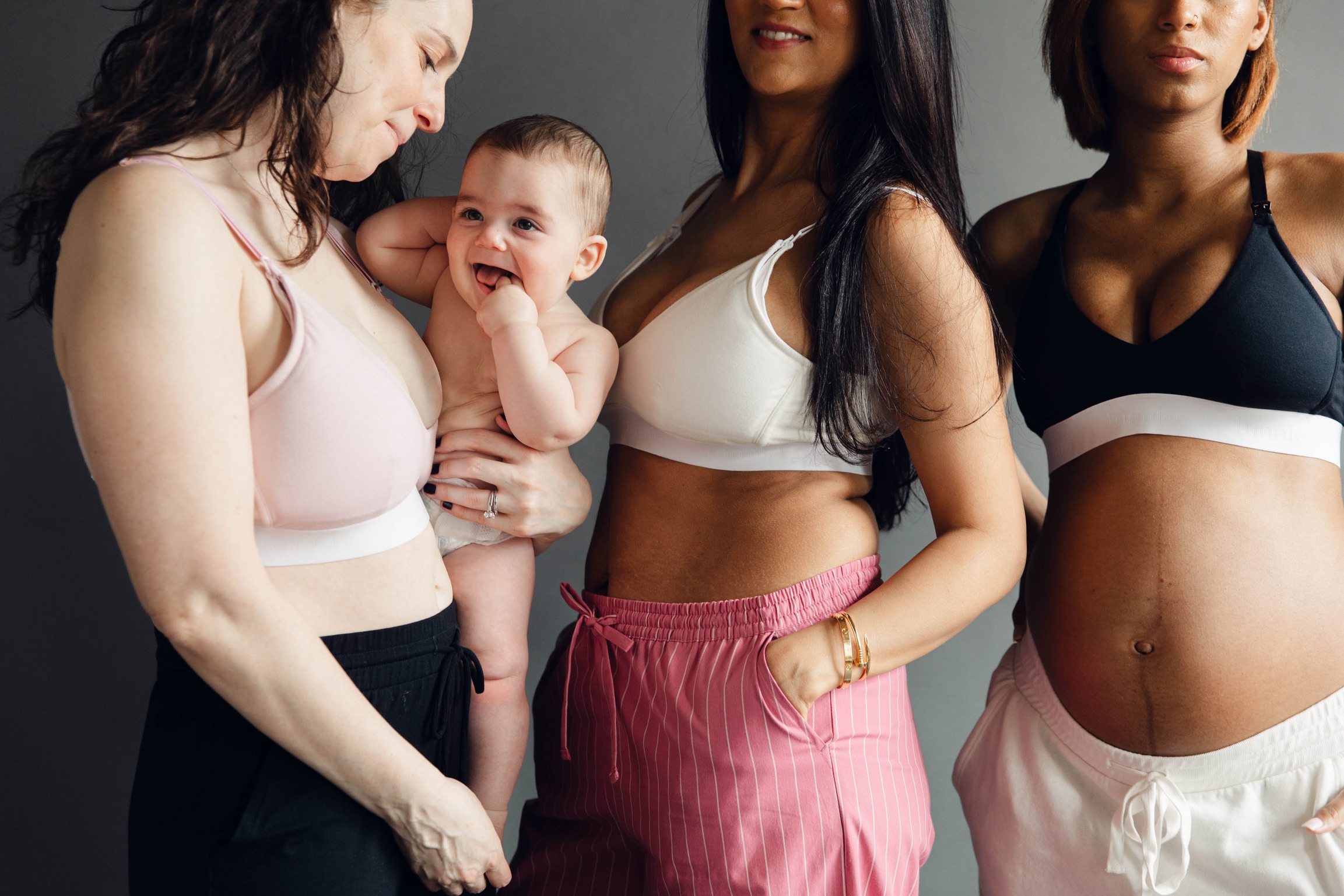 Four women and a baby pose together, with the women wearing sports bras and casual pants, smiling and looking at each other.