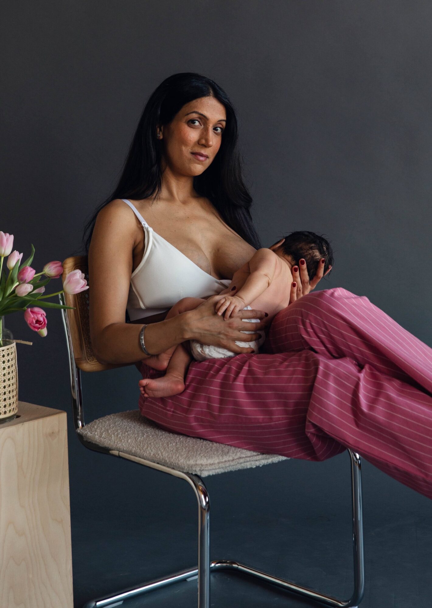 Woman in white top breastfeeding a baby, sitting on a chair with pink pants, against a dark background.