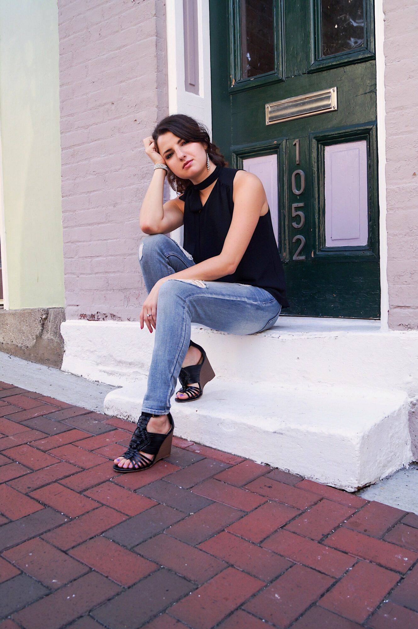 Young woman sitting on steps outside a building, resting her head on her hand, wearing a black top and jeans.