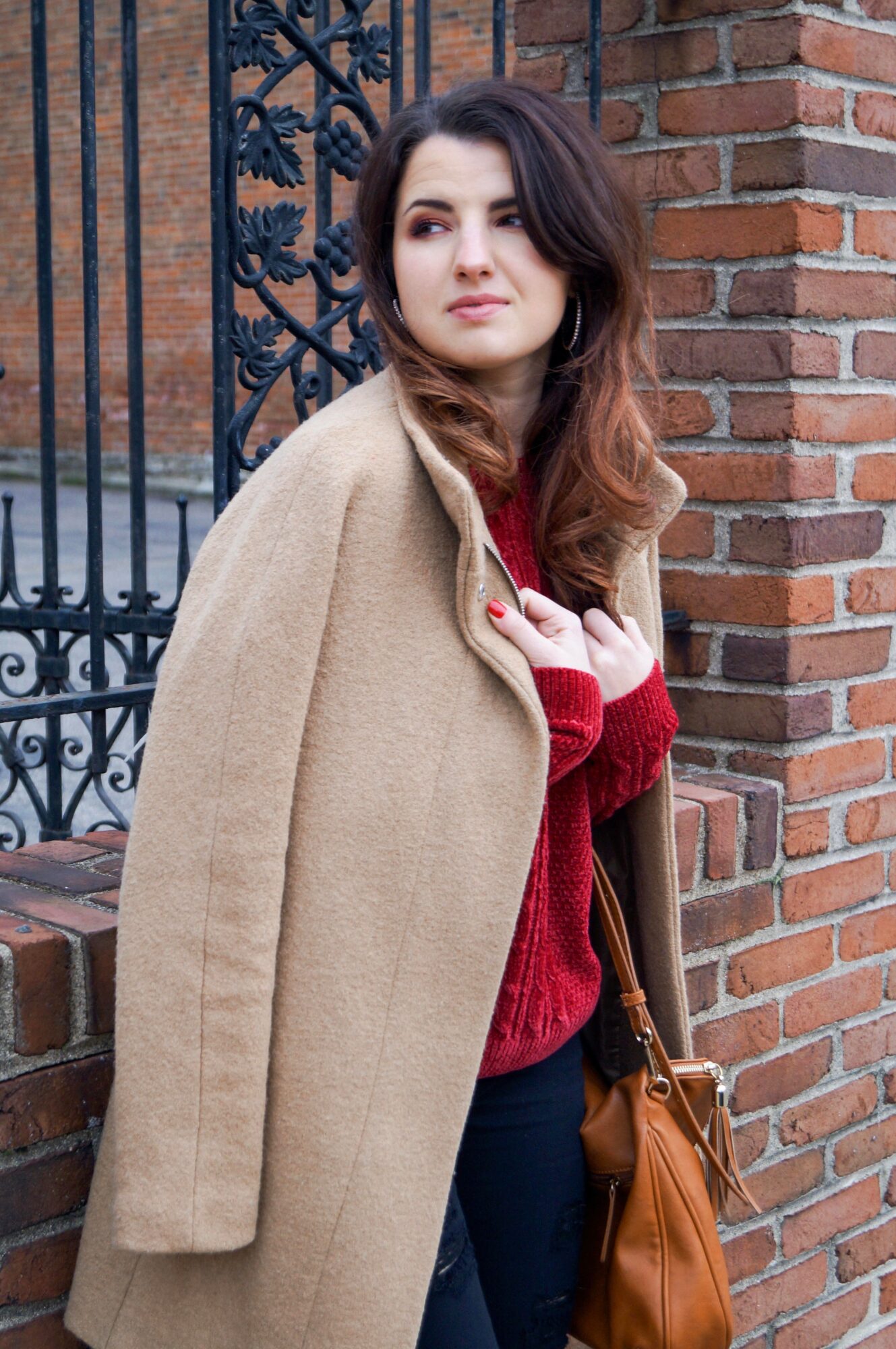 Young woman with wavy brown hair stands near a brick wall and black iron fence, wearing a beige coat and red sweater.