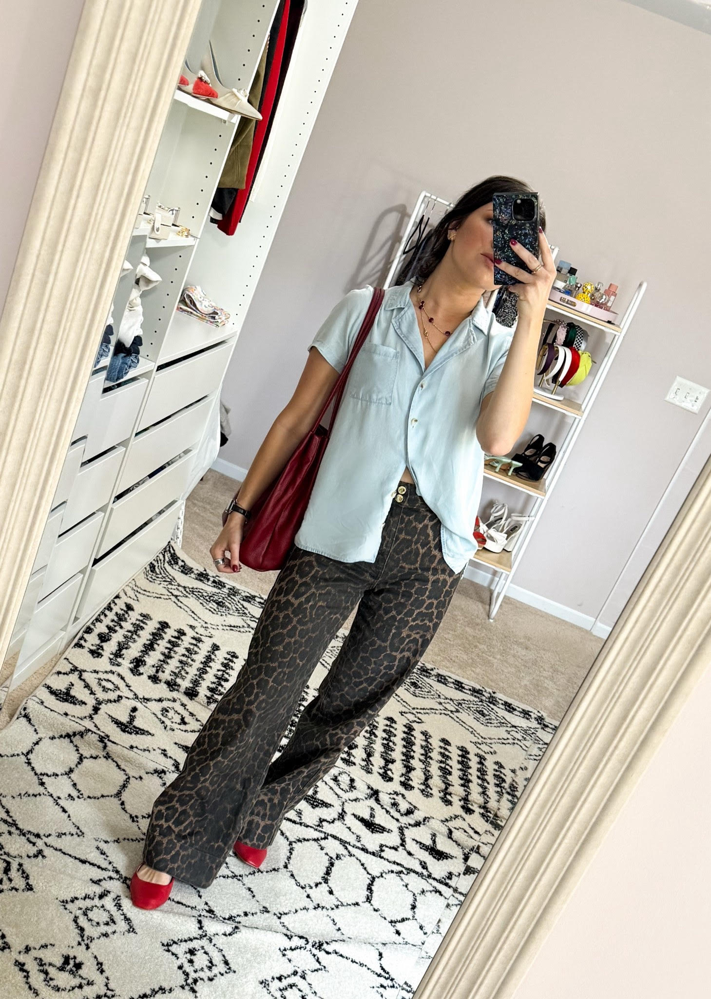 Person taking a mirror selfie in a room with shelves, wearing a light blue shirt, patterned pants, and red shoes, carrying a red bag.