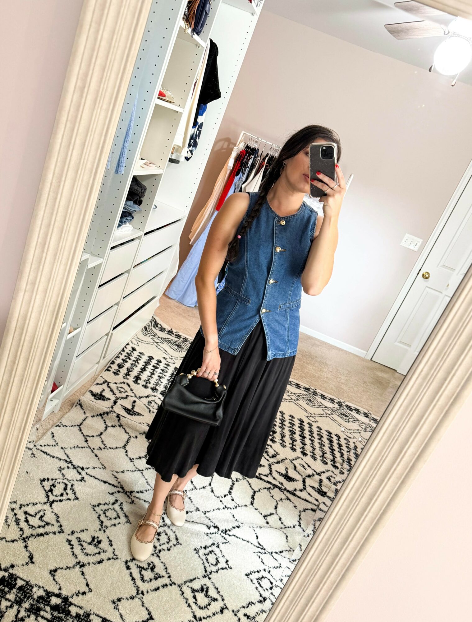 Woman taking mirror selfie in a room with white walls, wearing a blue sleeveless top, black skirt, and beige shoes, holding a black bag.
