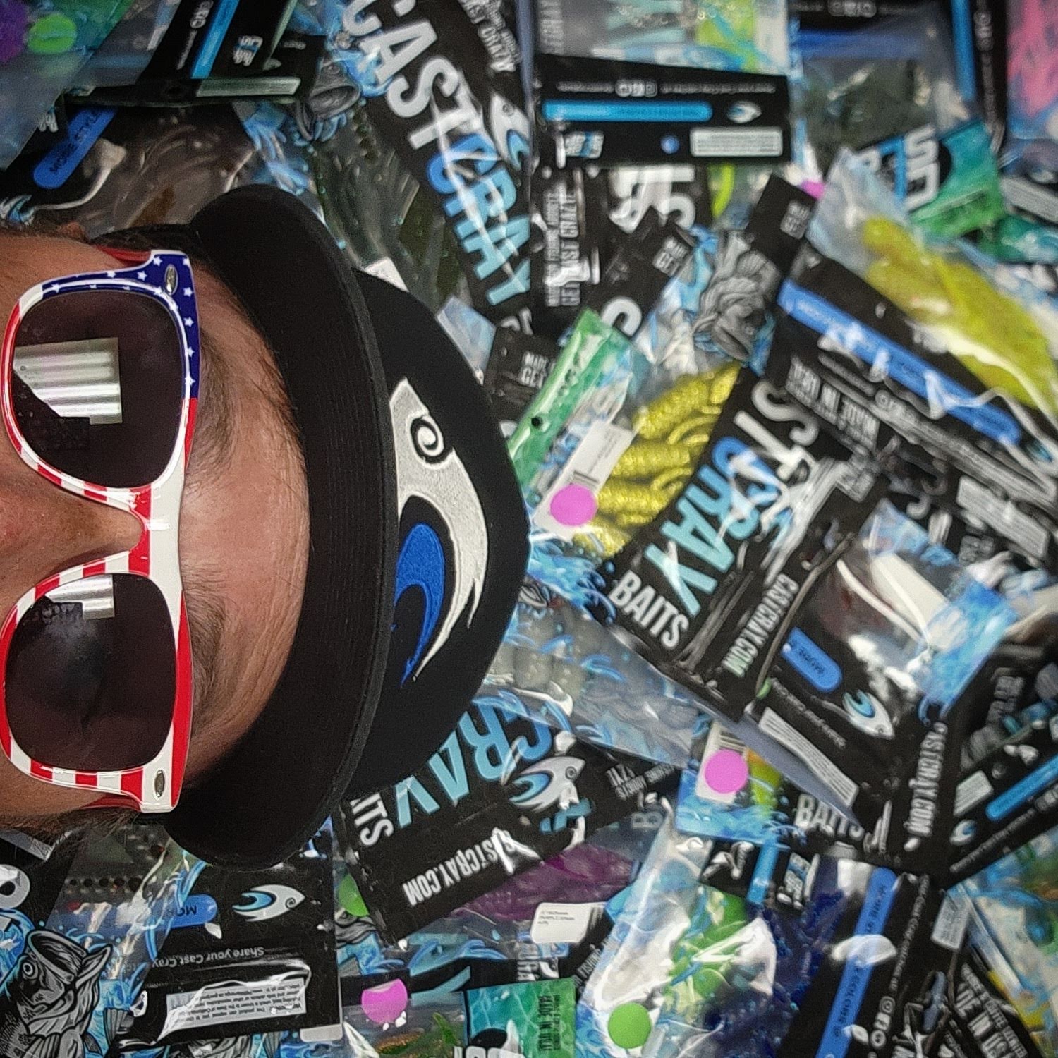 Person wearing patriotic sunglasses and a black cap surrounded by fishing bait packages.