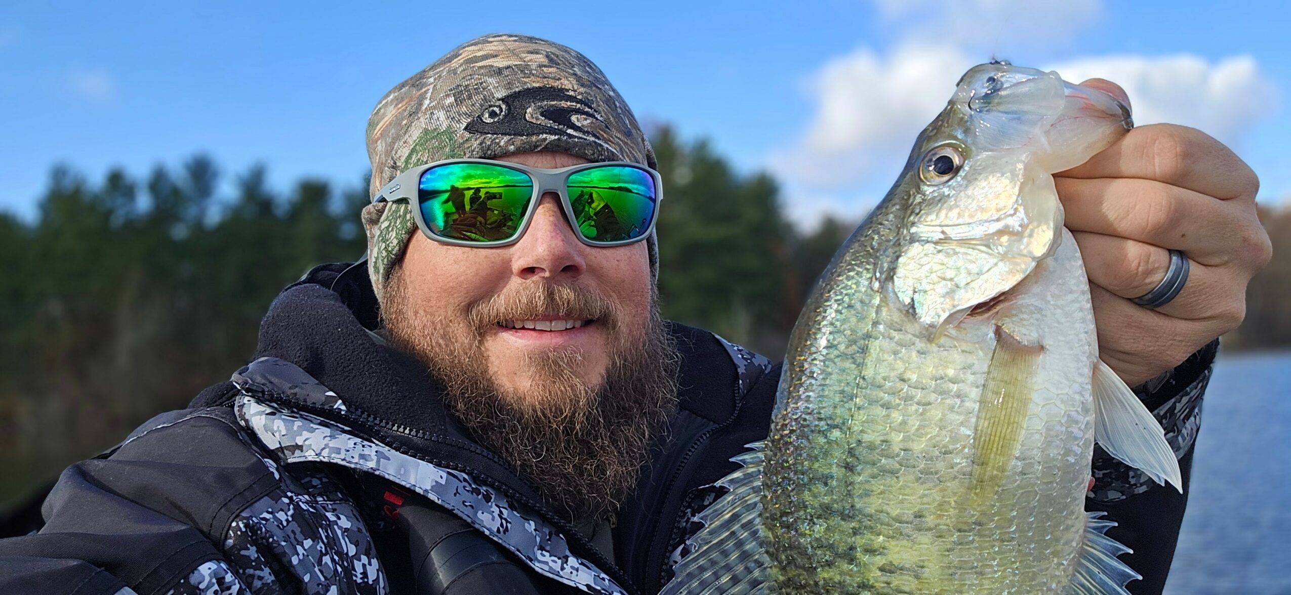 Man with sunglasses and a beard holds a large fish outdoors, with trees and blue sky in the background.
