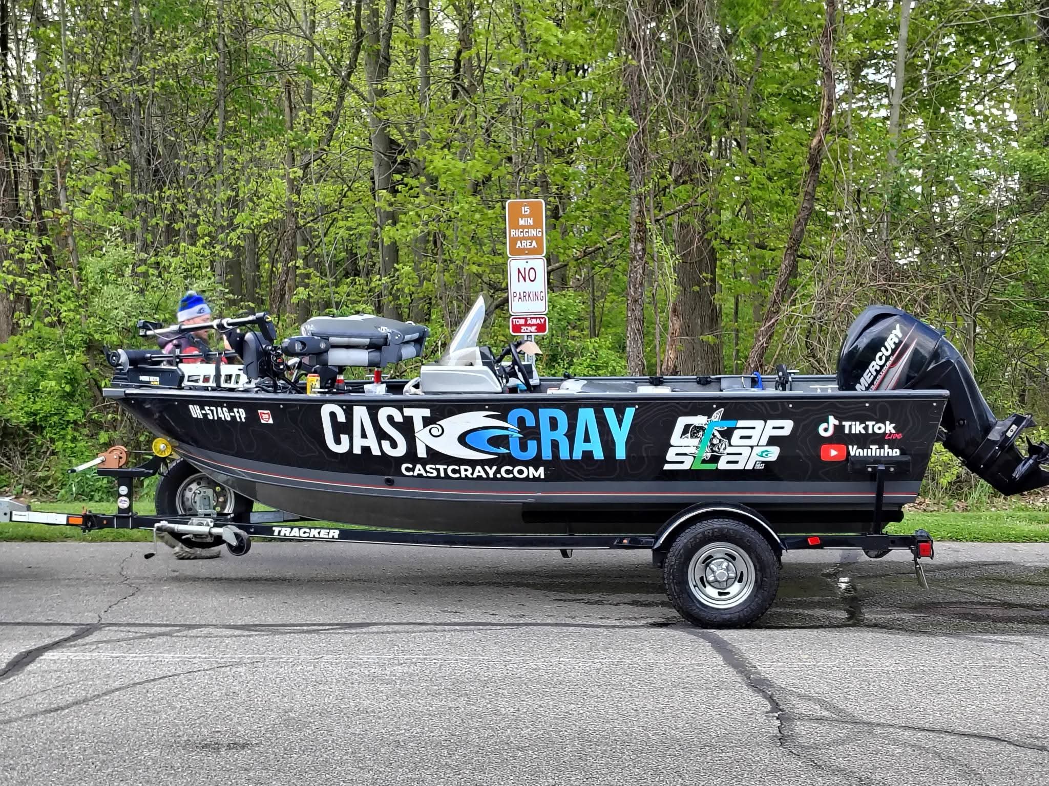 A black boat on a trailer parked on a street with trees in the background, displaying various logos and a motor at the back.