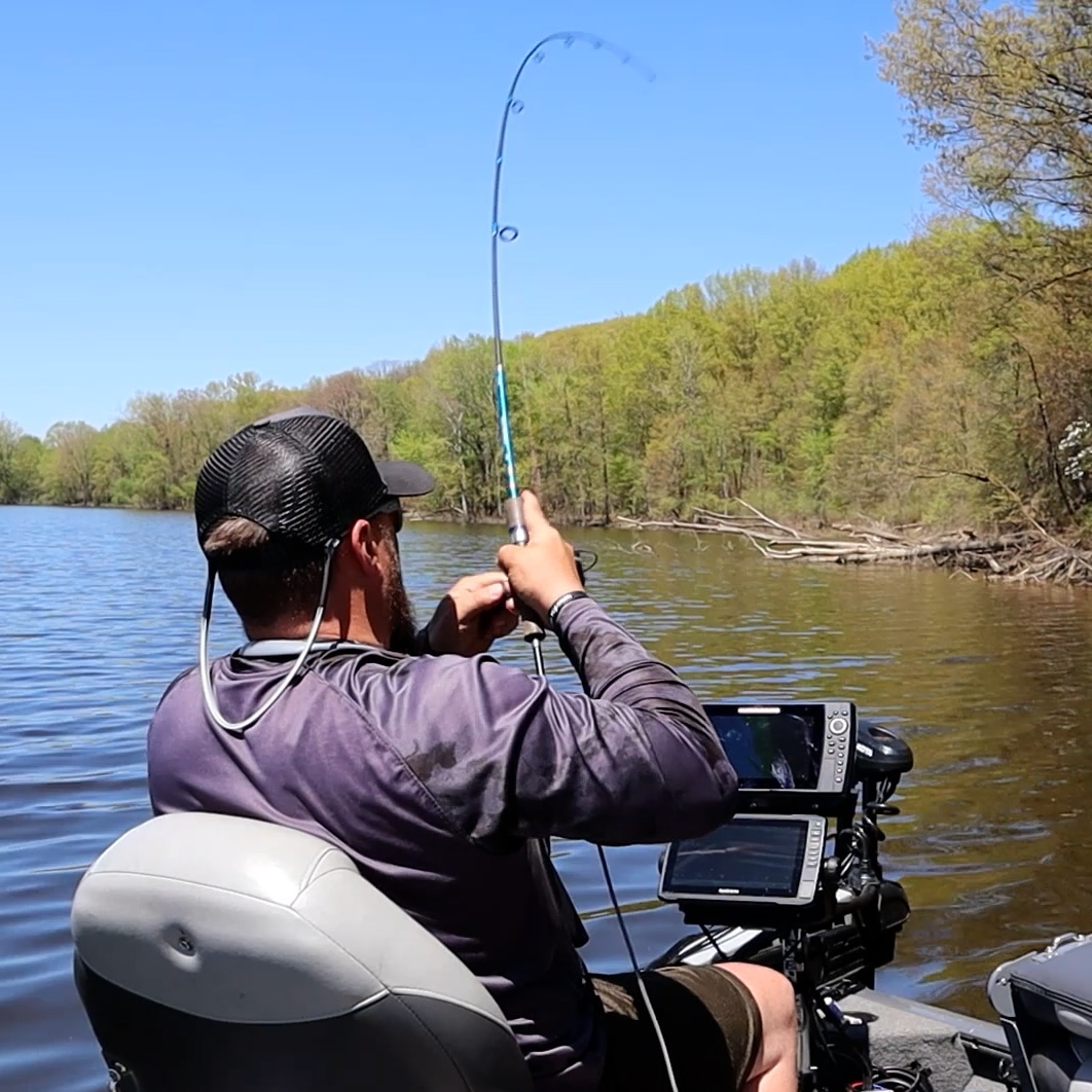 Person fishing on a boat in a river with trees in the background, holding a fishing rod, wearing a cap and jacket.