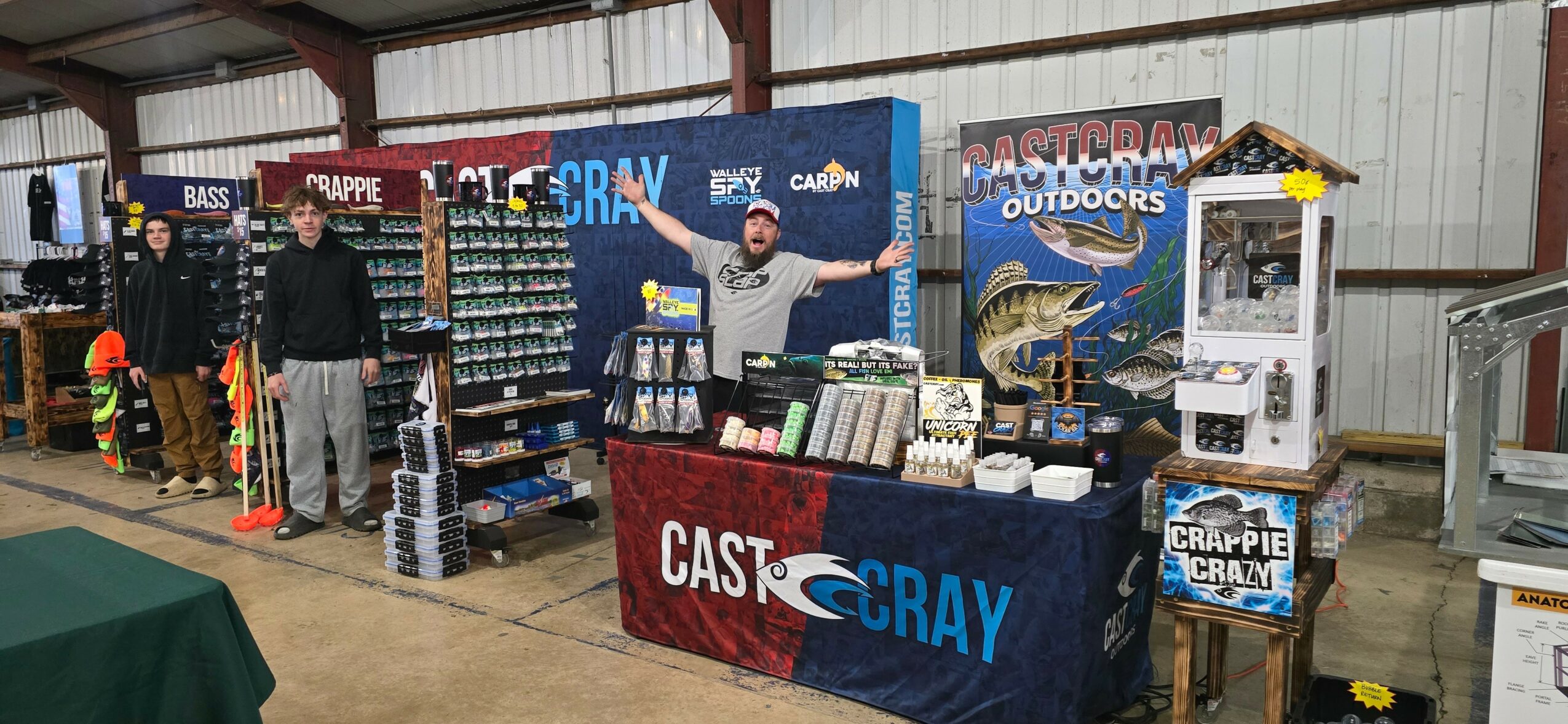 Person standing behind a booth with fishing gear and a large fish display, inside a warehouse-like space.