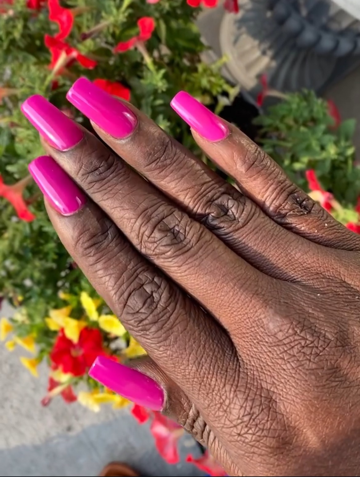 Hand with pink nail polish resting on colorful flowers, showing detailed skin texture and long nails.