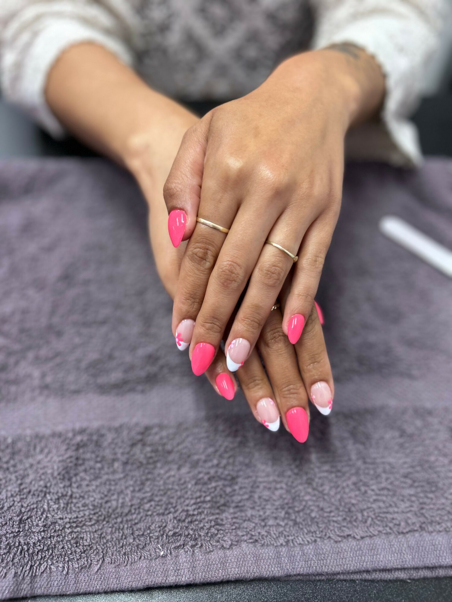 Hands with pink and white nail polish resting on a gray towel, wearing rings, with a blurred background.