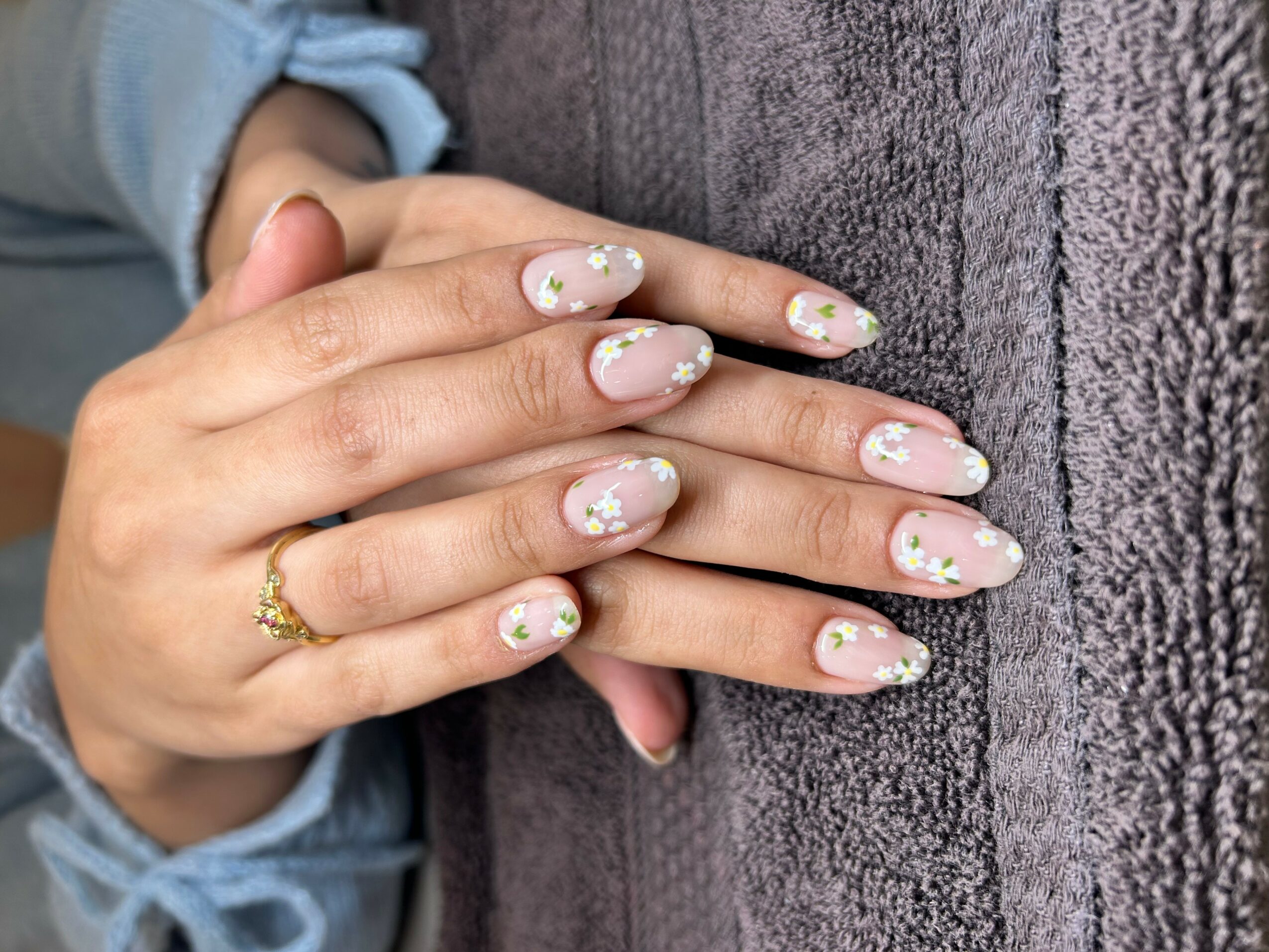 Person's hand with decorated nails resting on textured gray surface, wearing a gold ring and light blue sleeve, nails with floral design