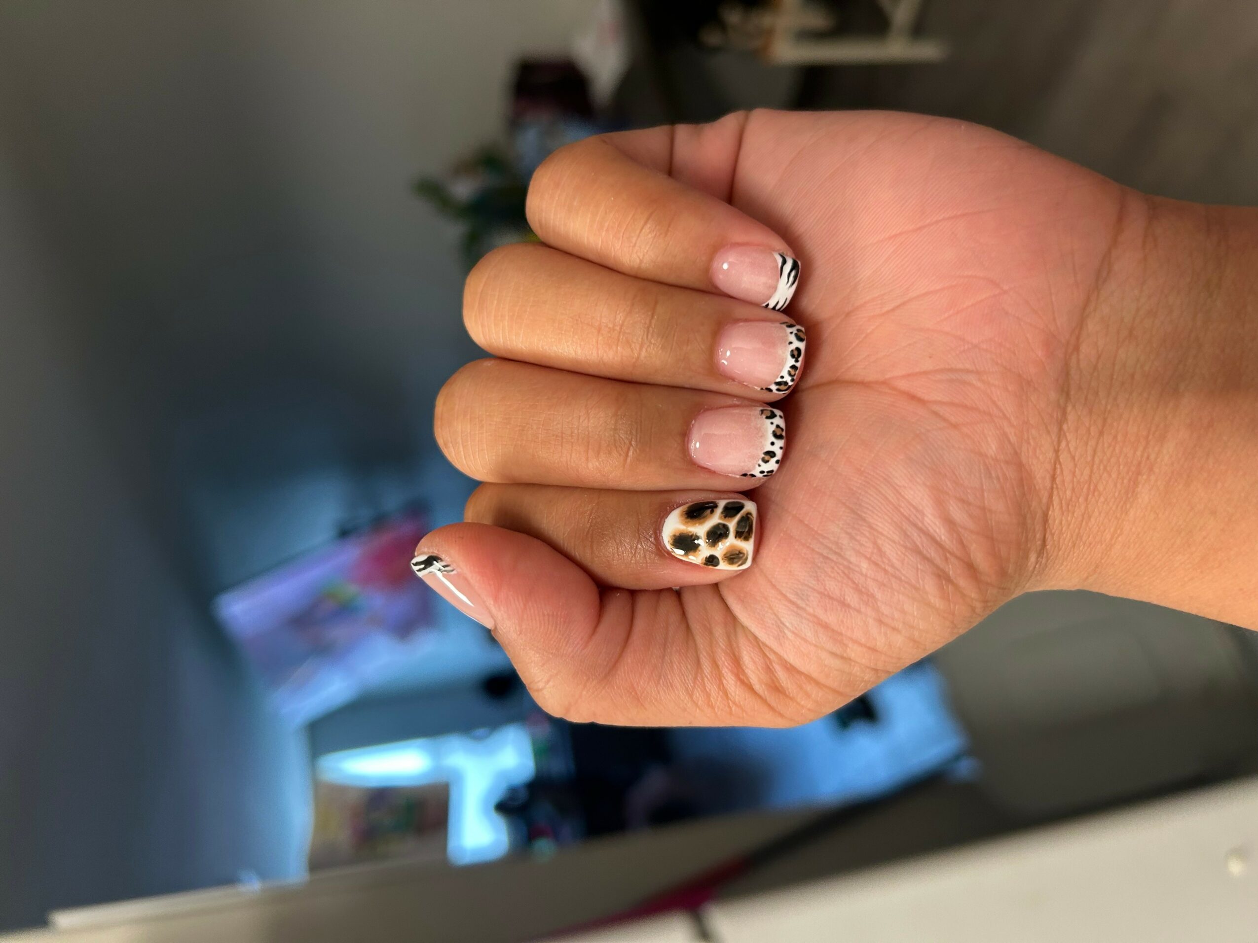 Hand with decorated nails, some with black and white patterns, one with leopard print, in a room with blurred background.