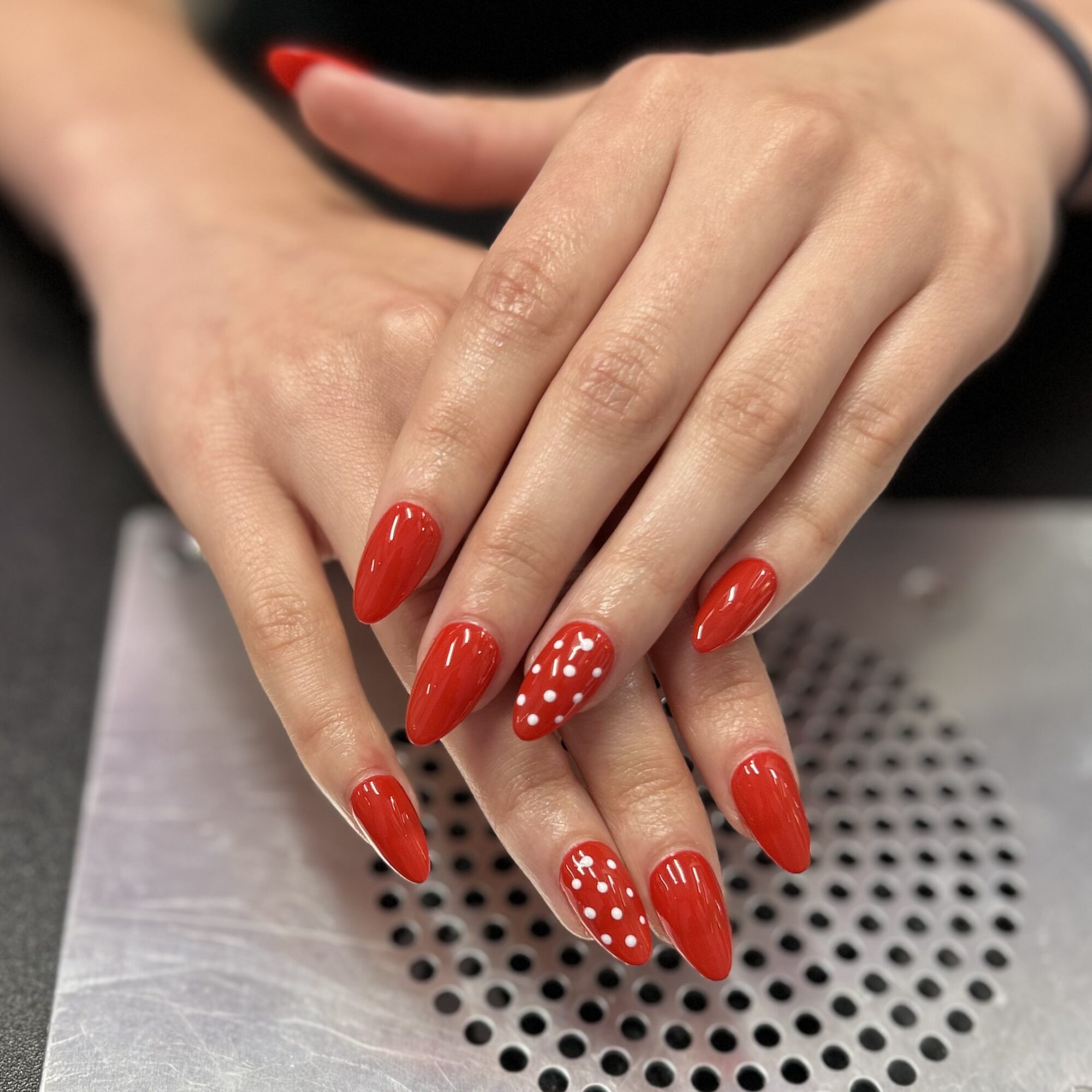 Hands with red nail polish and white polka dots on some nails, resting on a metallic surface.