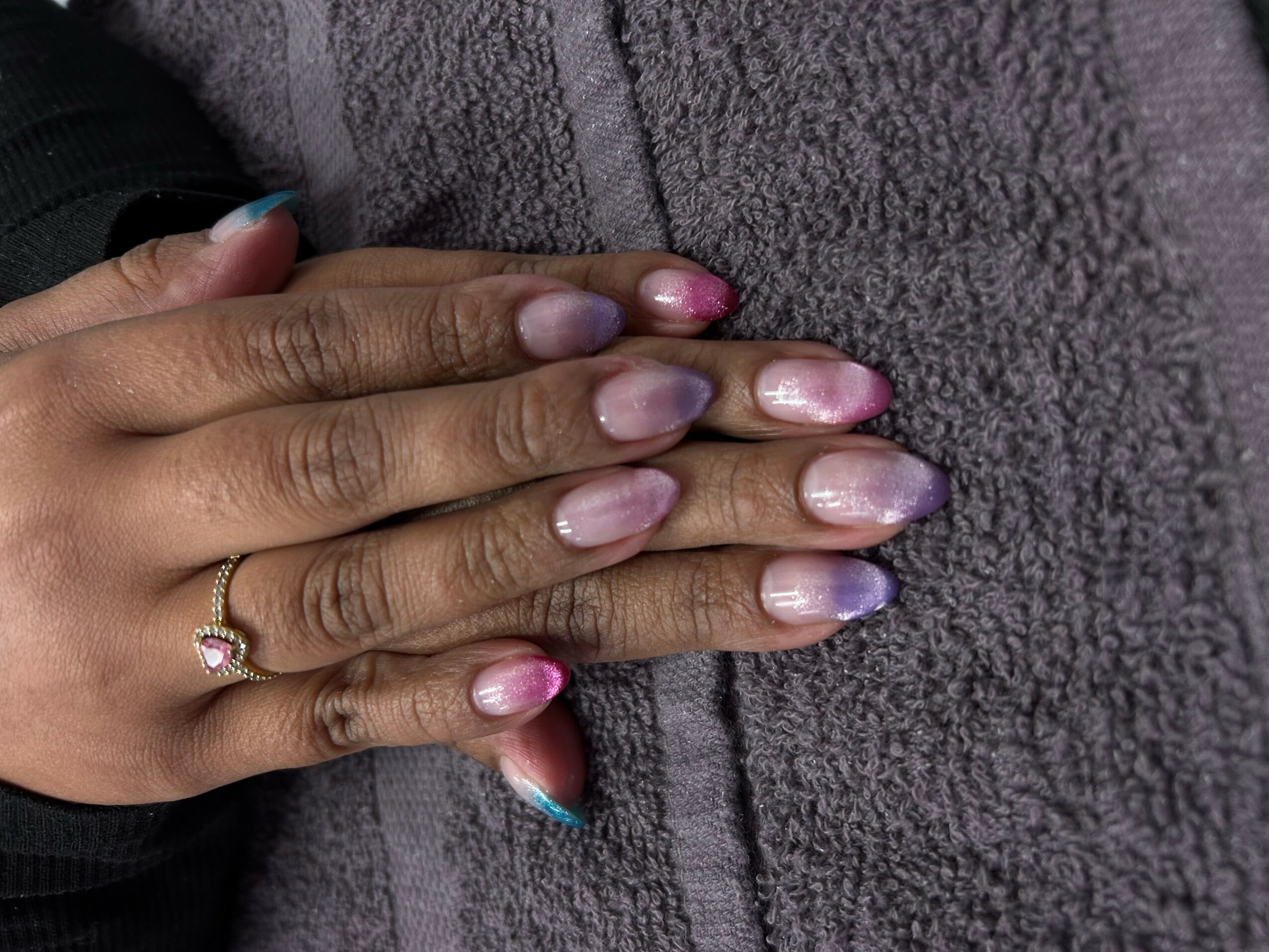 Two hands with manicured nails resting on a gray textured surface, one hand wearing a ring with a small gemstone.