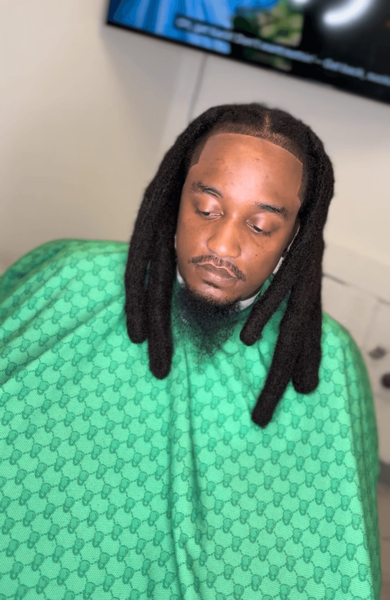 Man with long dreadlocks wearing a green patterned shirt, looking down, in an indoor setting with a TV screen in the background.