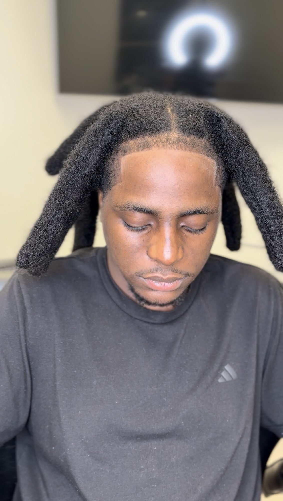 Young man with closed eyes and natural textured hair, wearing a black Adidas shirt, indoors with a blurred screen in background.