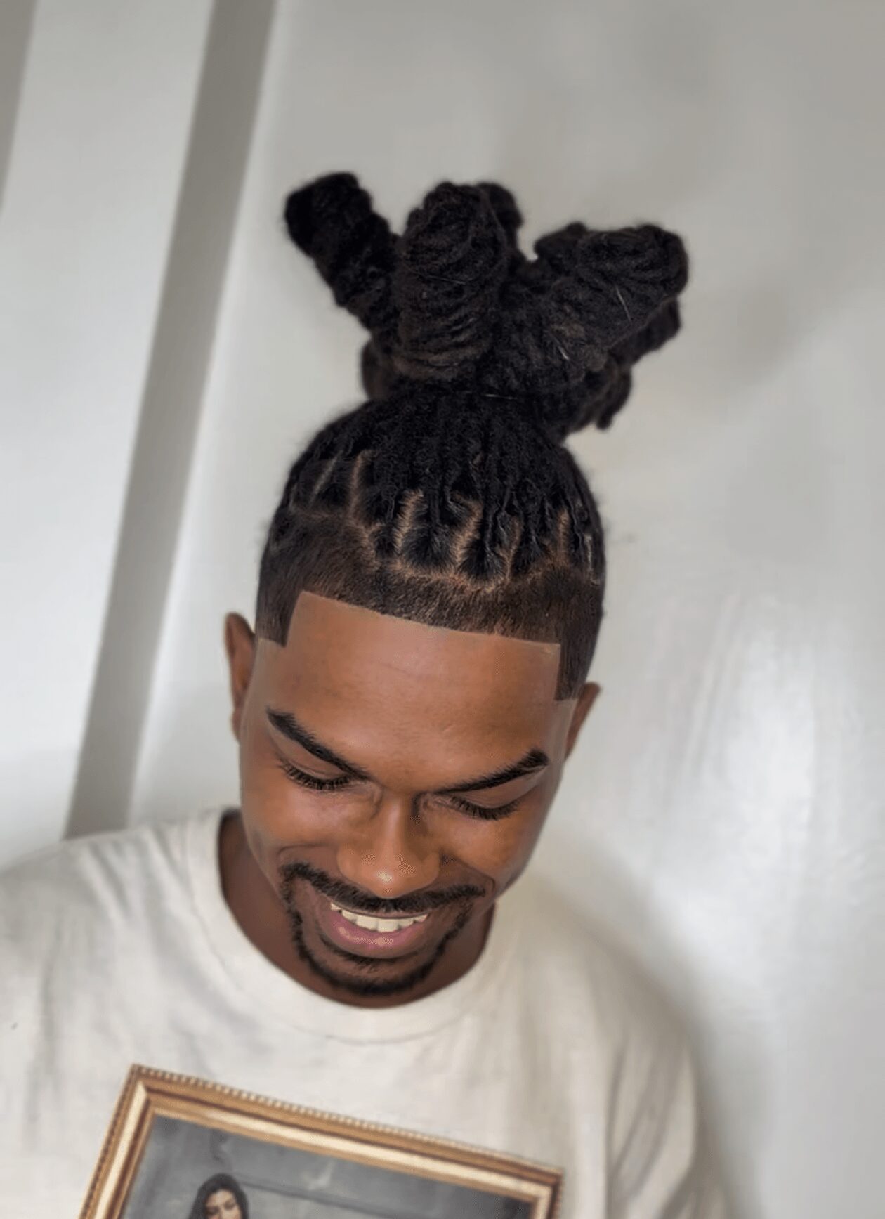 Young man with styled dreadlocks tied in a bun, smiling, wearing a white shirt, against a plain background.