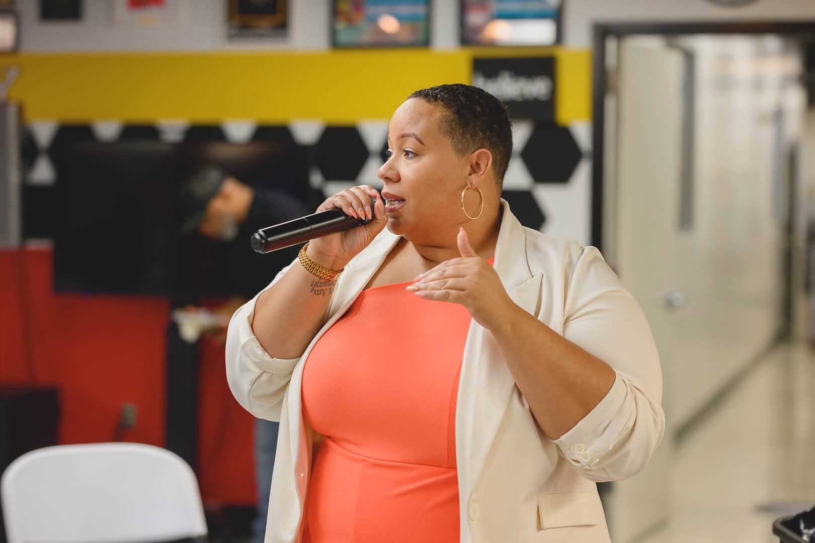 Woman singing into a microphone indoors, wearing a coral dress and cream blazer, with a yellow wall in the background.