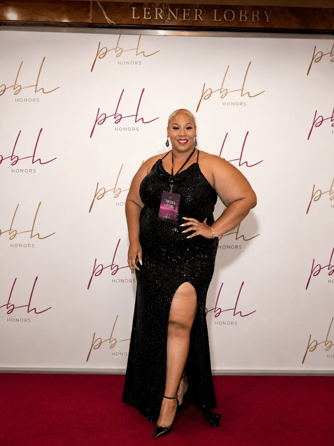 Woman in a black gown with a thigh-high slit, smiling, standing on a red carpet in front of a backdrop with 'pbh' logos.