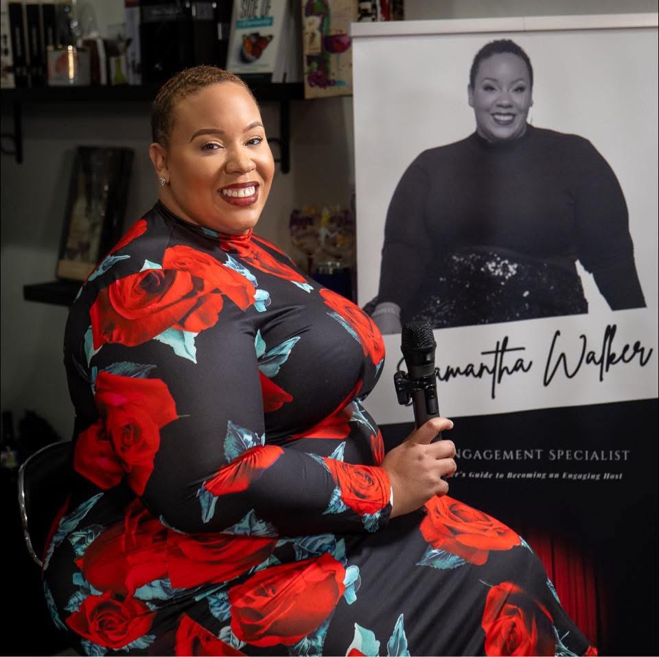 Smiling woman in floral dress holding a microphone, sitting next to a large black-and-white portrait of herself.