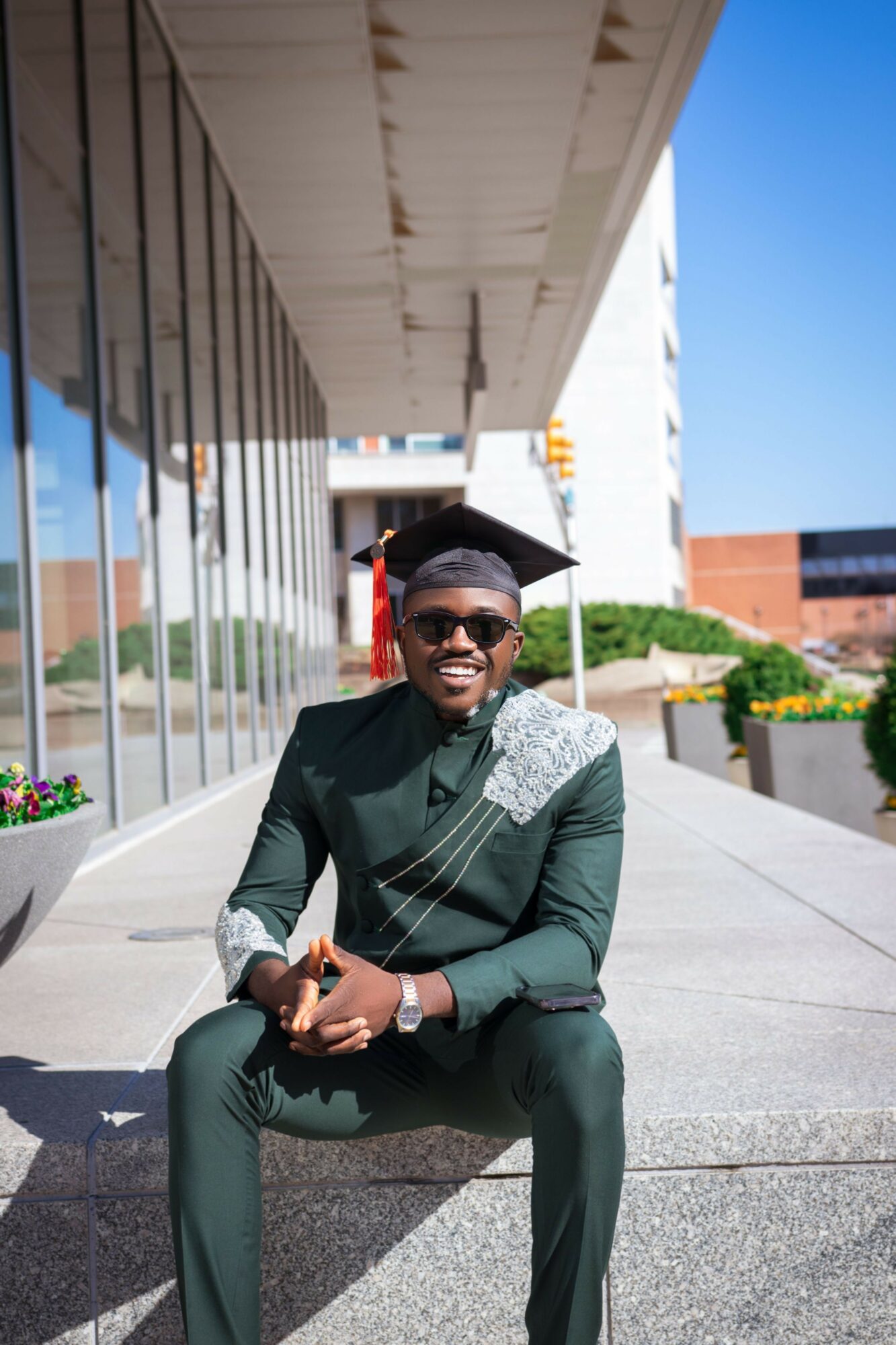 Person in graduation cap and gown sitting outdoors on a sunny day, smiling, with modern building and flowers in background.