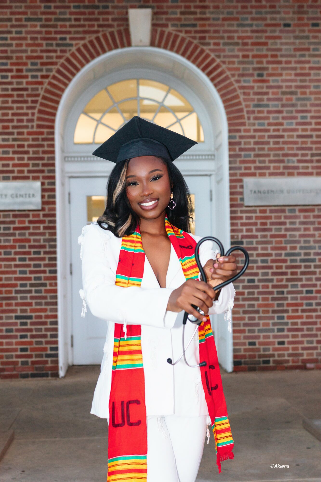 Graduate in white suit with colorful stole and cap, holding a heart-shaped award, standing in front of a brick building with an arched window.