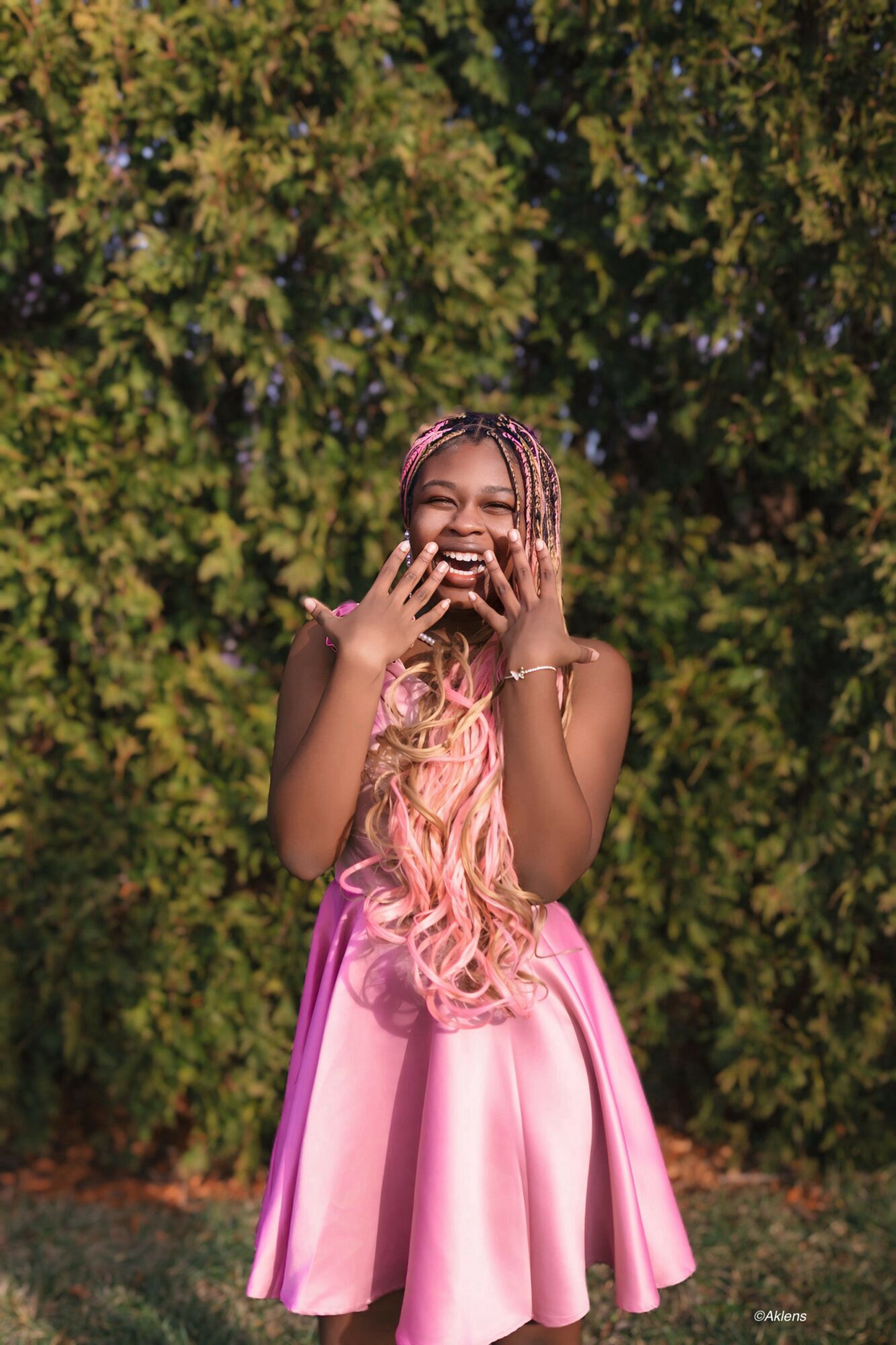 Young woman with long pink hair laughing outdoors, wearing a pink dress, standing in front of green trees.