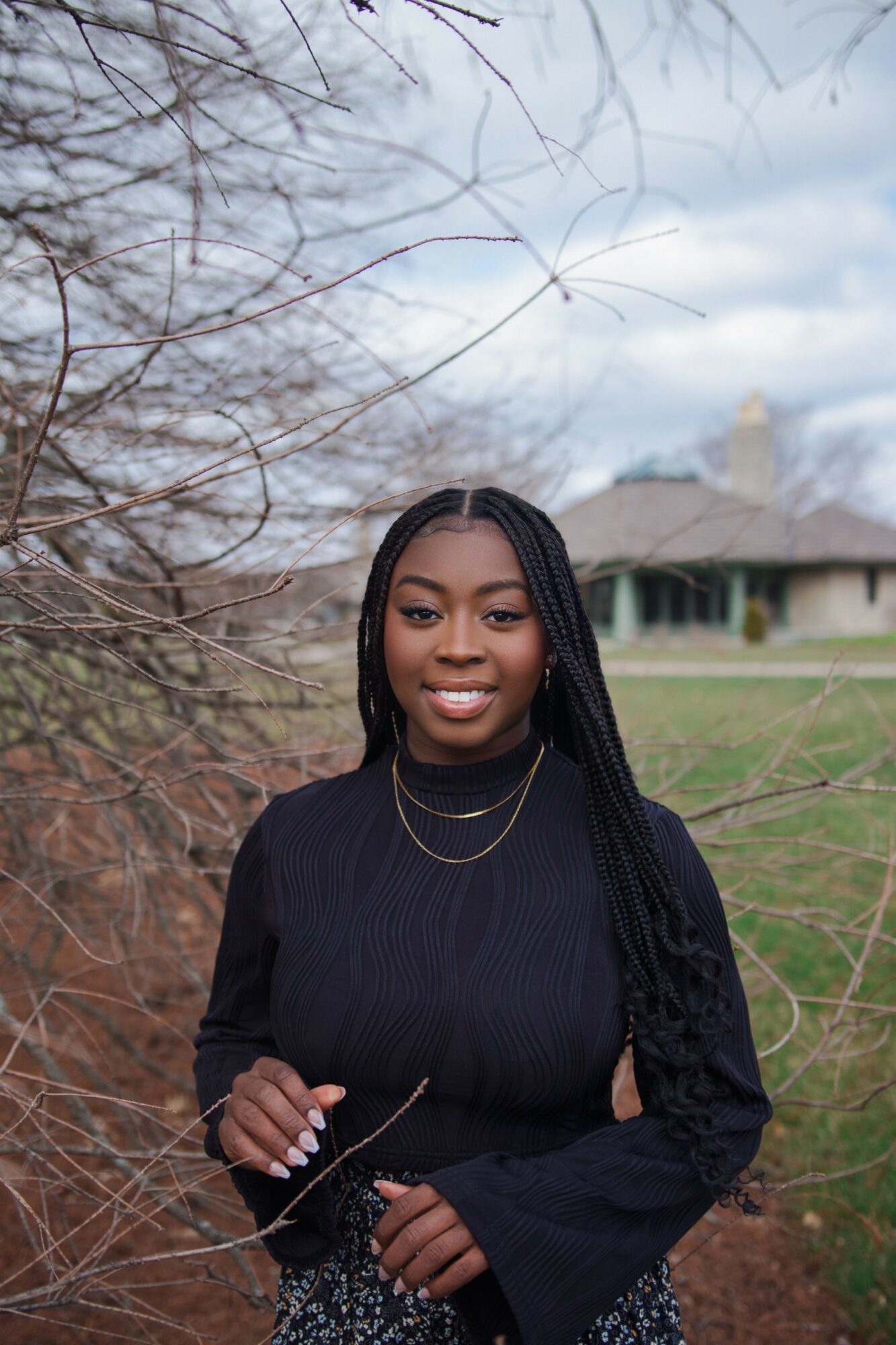 Young woman with long braided hair smiling outdoors, wearing a black sweater and layered necklaces, with trees and a house in background.