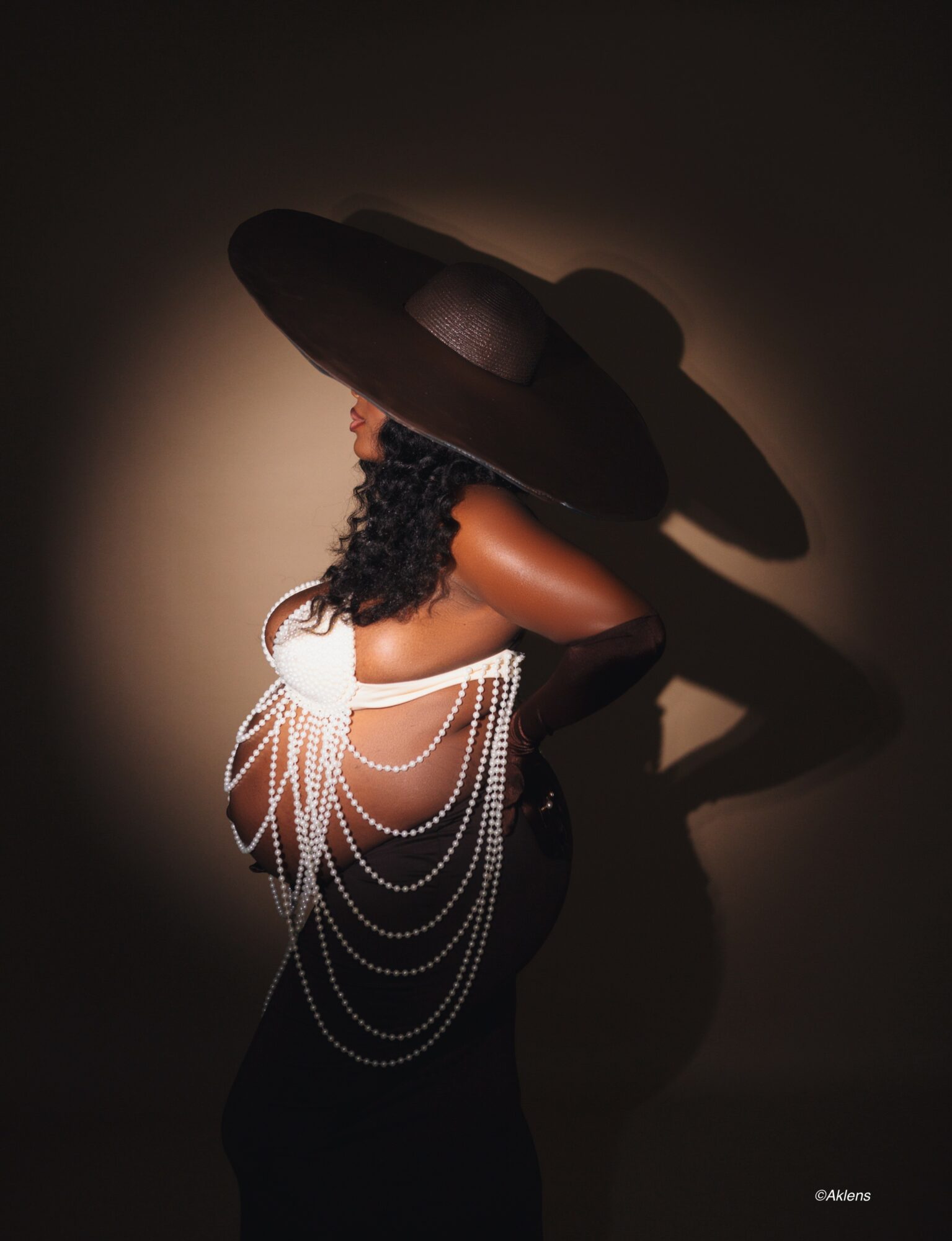 Woman wearing a large hat with pearl accessories, facing sideways, shadow cast on wall, dark background.