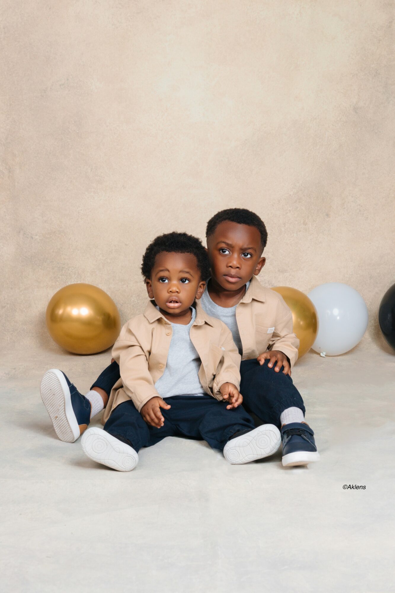 Two young children sit on the floor with balloons behind them, one child is slightly older and both wear beige jackets.