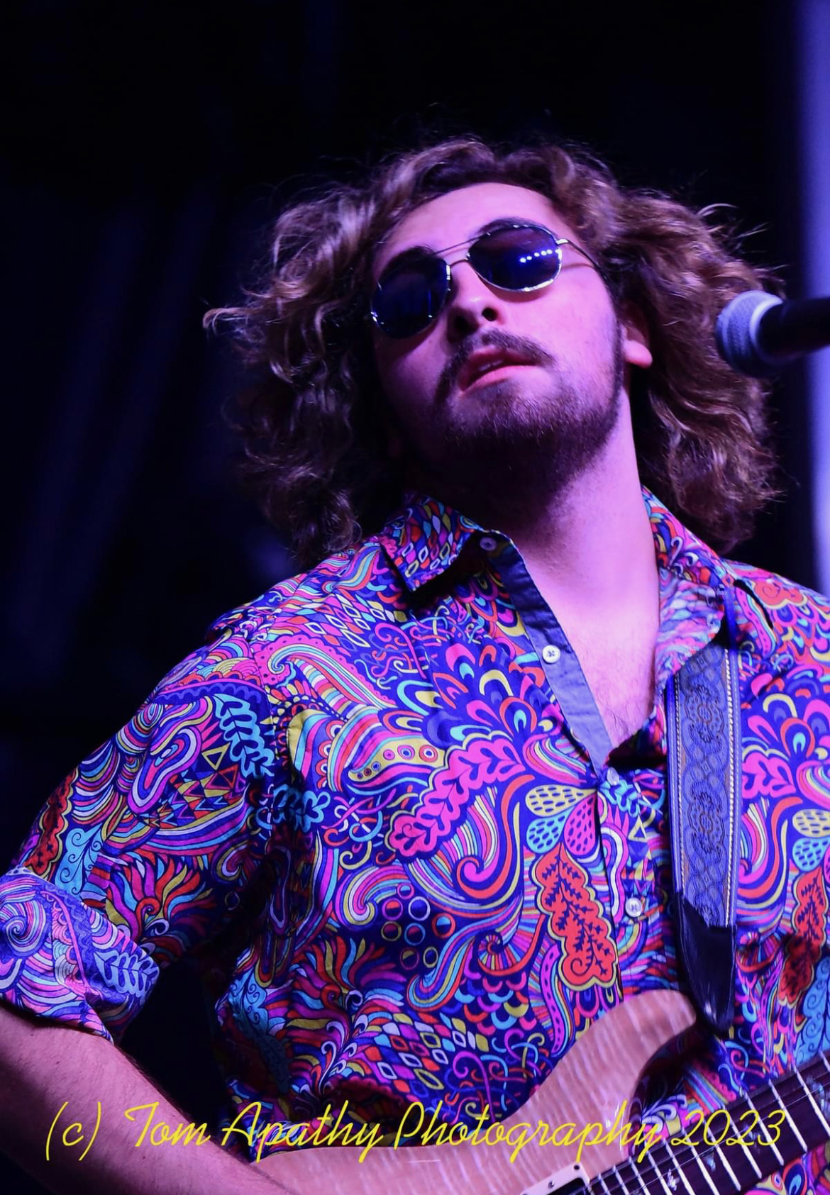 Person with curly hair wearing sunglasses and a colorful patterned shirt, standing near a microphone on stage.