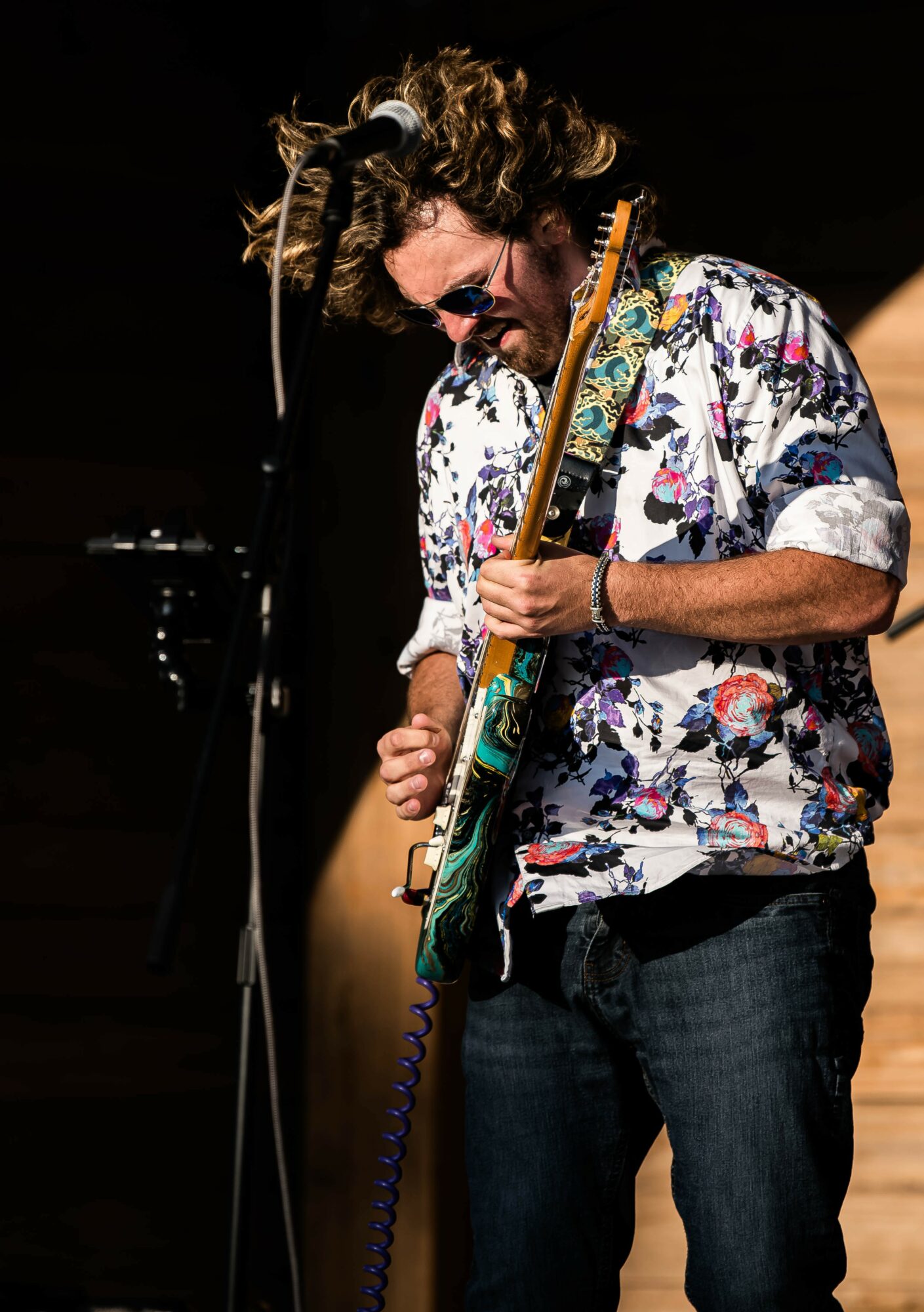 Person playing guitar, wearing sunglasses and a floral shirt, with curly hair, on stage in a dark setting.