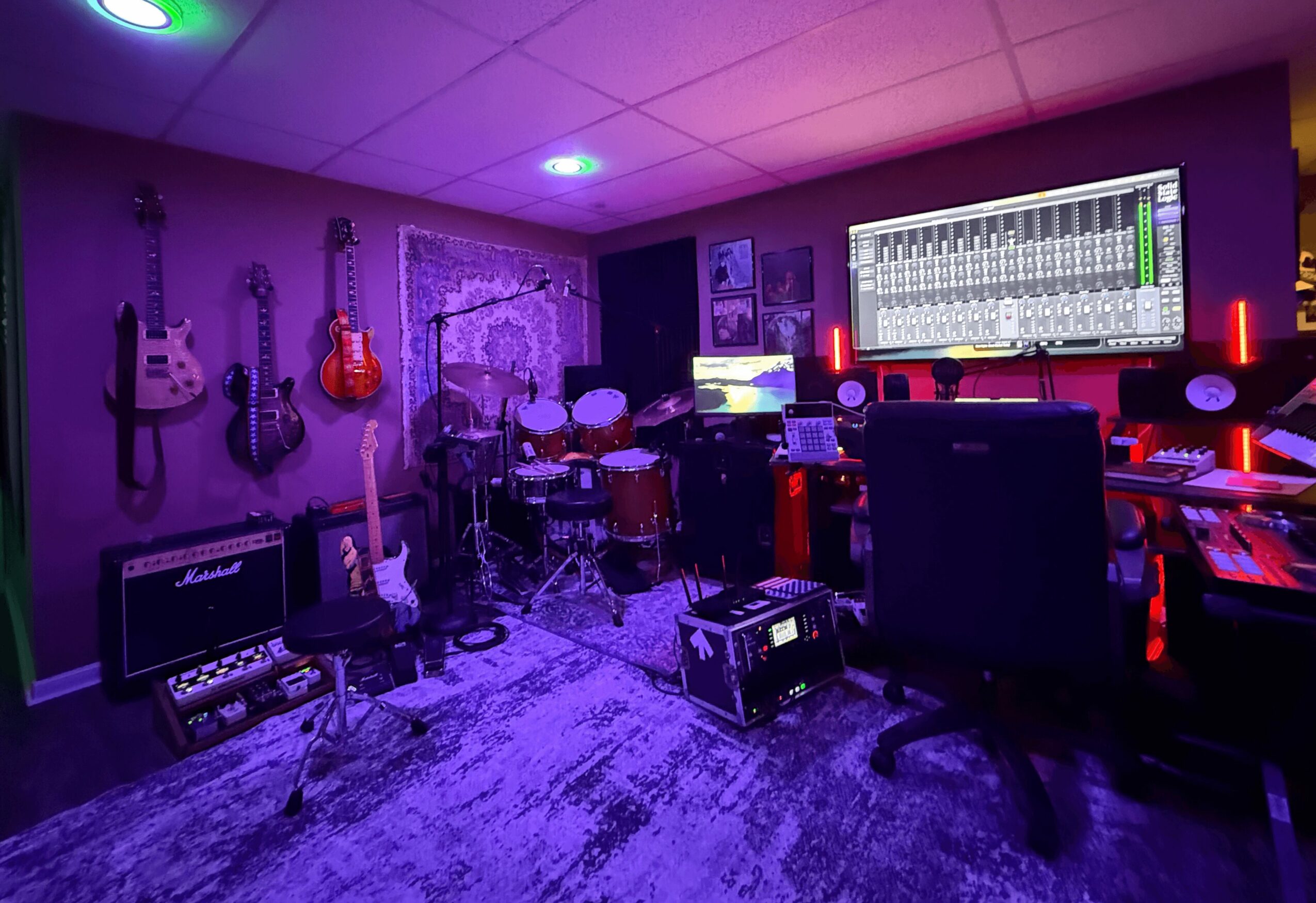 Music studio with guitars, drum set, mixing console, and computer screens, illuminated by colorful LED lights.